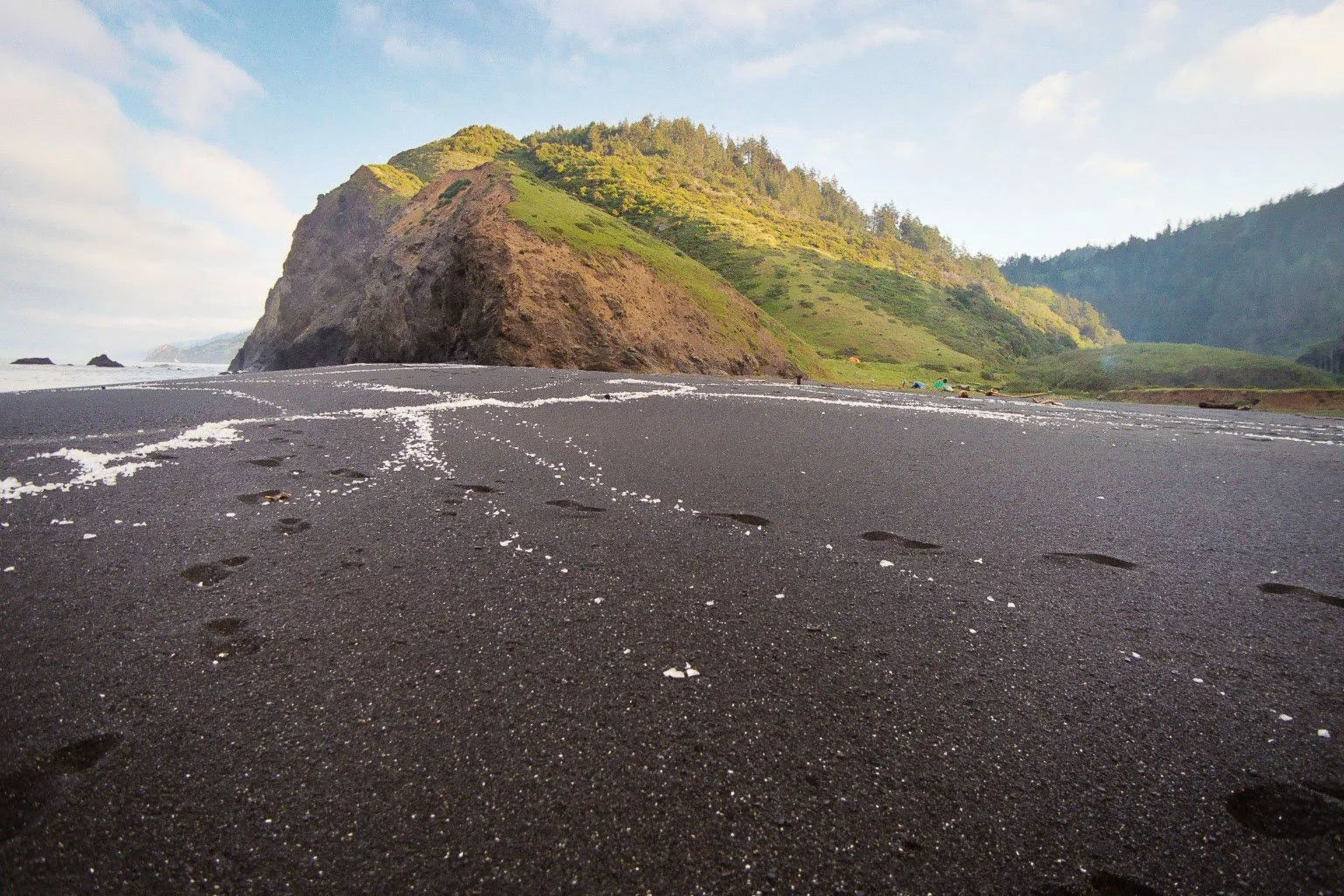 A black sand beach with mountain scenery in the background and footprints in the sand.