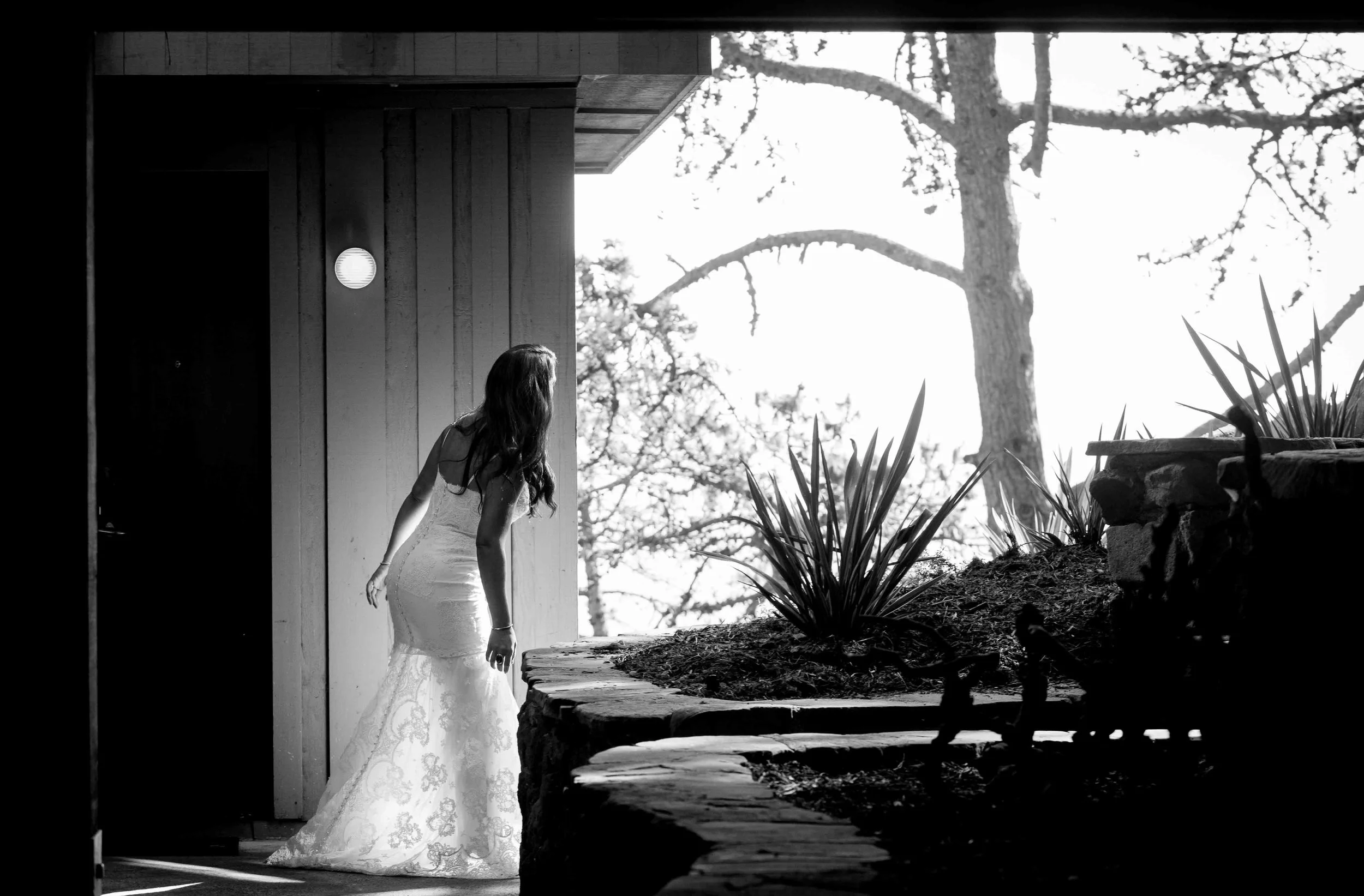 A woman in a wedding dress is looking down, standing outside near a wooden structure and plants, with trees in the background.