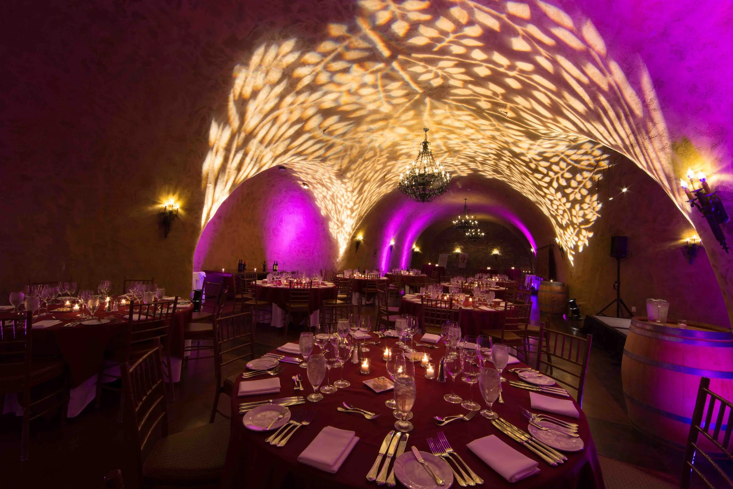 Elegant banquet hall with vaulted ceilings, illuminated with purple and gold lighting, set with round tables covered with burgundy tablecloths, fine china, glassware, and lit candles, decorated for a formal event.