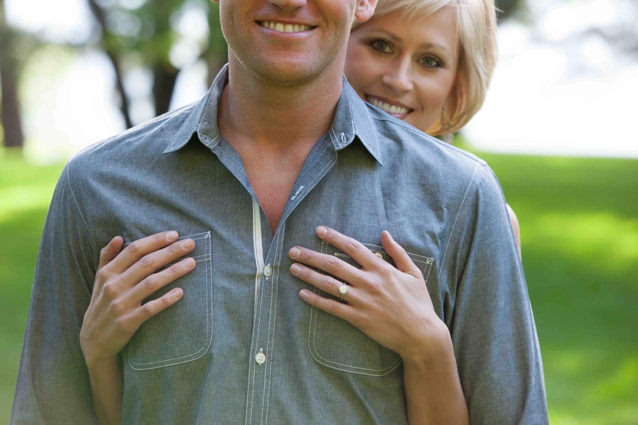 A couple stands outdoors in a park, with the woman’s hands resting on the man’s chest, showing an engagement ring. The woman is smiling behind the man, who is also smiling. Green trees and grass are in the background.