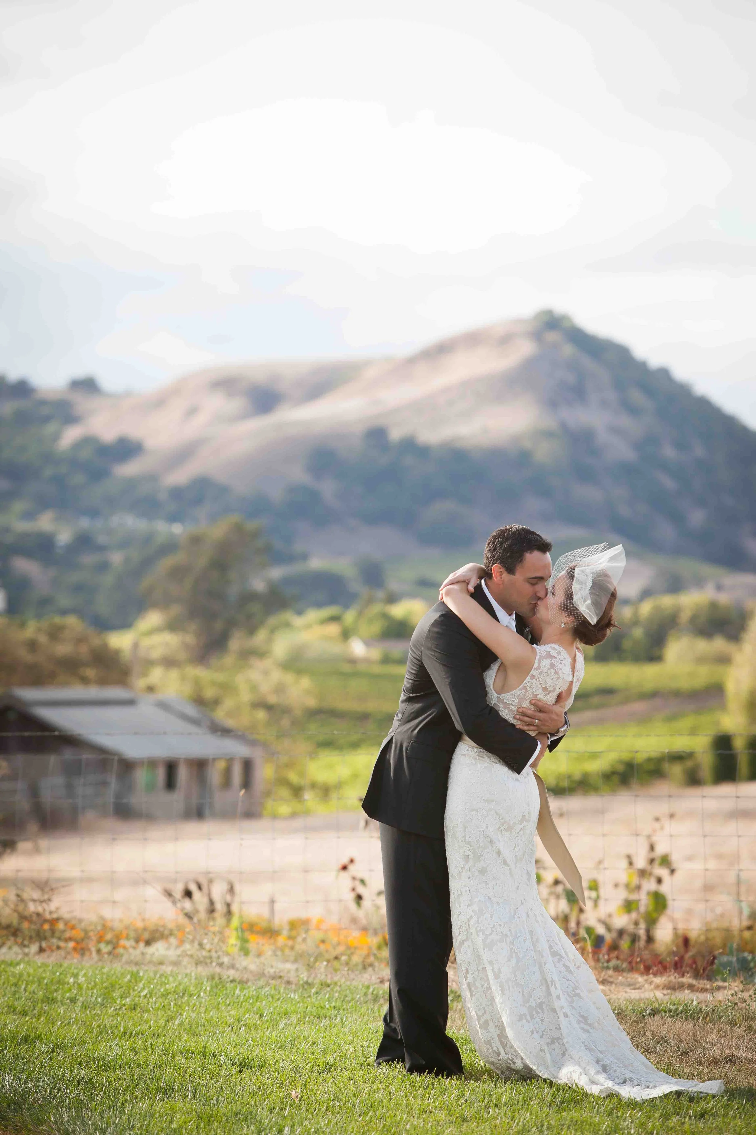 A bride and groom sharing a kiss outdoors on a grassy field with mountains in the background.