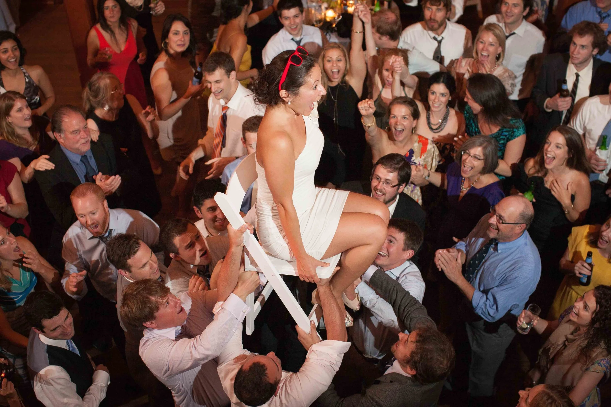 A woman in a white dress is sitting on a chair being lifted by multiple people at a party or celebration, surrounded by smiling and cheering guests.