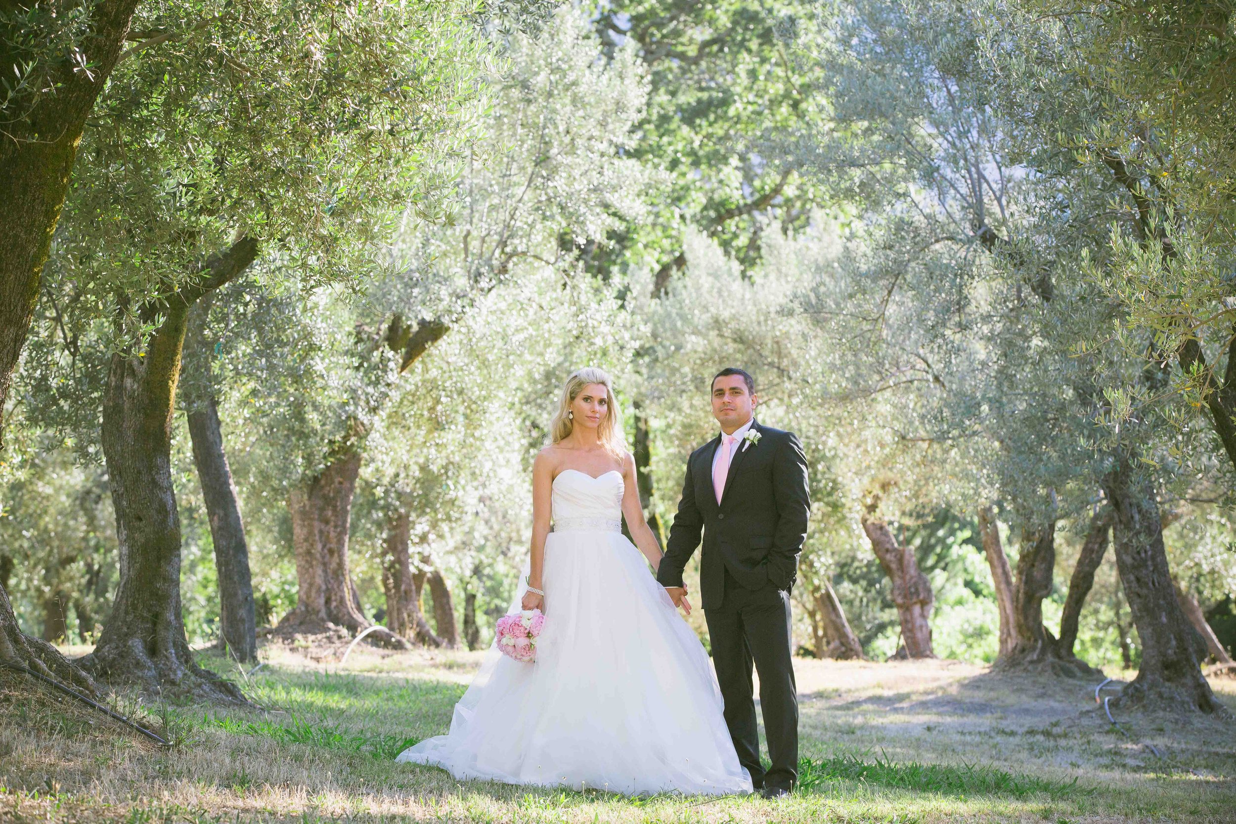 A bride in a white wedding gown holding a pink bouquet and a groom in a black suit standing hand in hand in a lush, green outdoor setting with trees and sunlight filtering through the leaves.