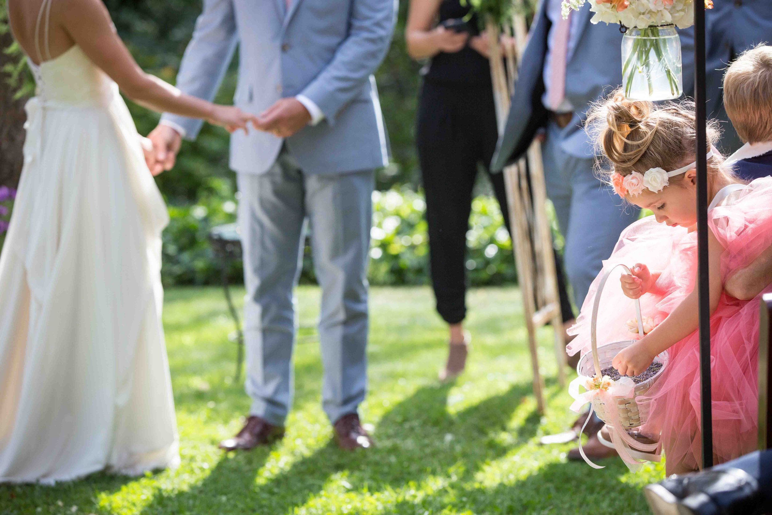 An outdoor wedding ceremony with a bride and groom holding hands, surrounded by guests, one of whom is a young girl in a pink dress picking flowers from a basket.