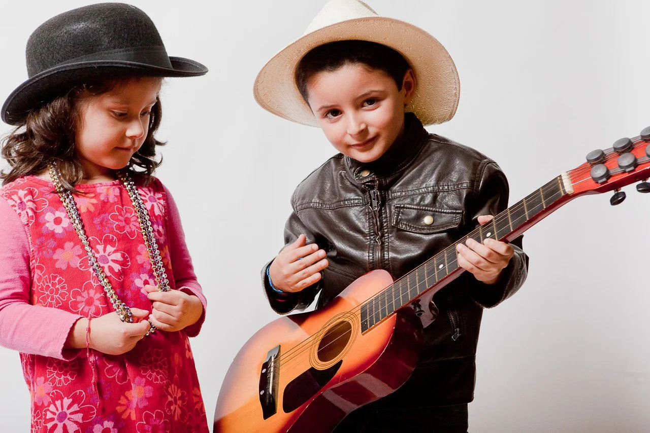 A young girl in a pink flower-patterned dress, wearing a black hat and necklaces, stands to the left. A young boy in a leather jacket and wide-brimmed hat, holding a guitar, stands to the right. He is smiling and looking at the camera.