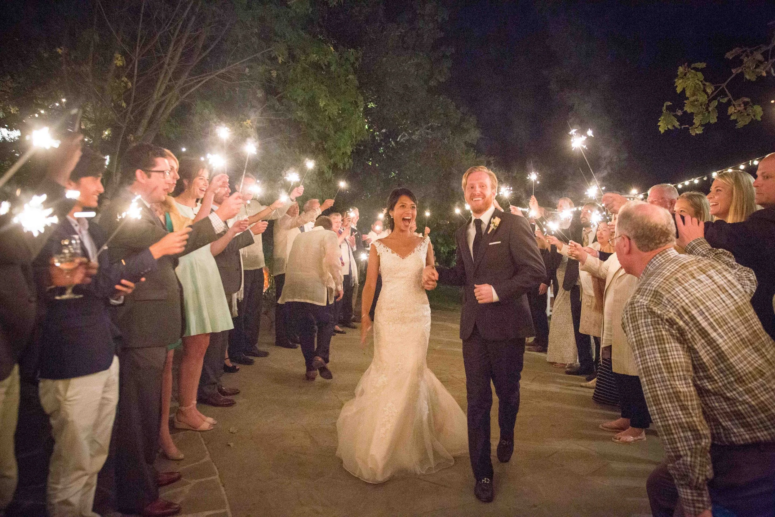 Bridal couple walking hand in hand through a candlelit outdoor celebration with guests holding sparklers at night.