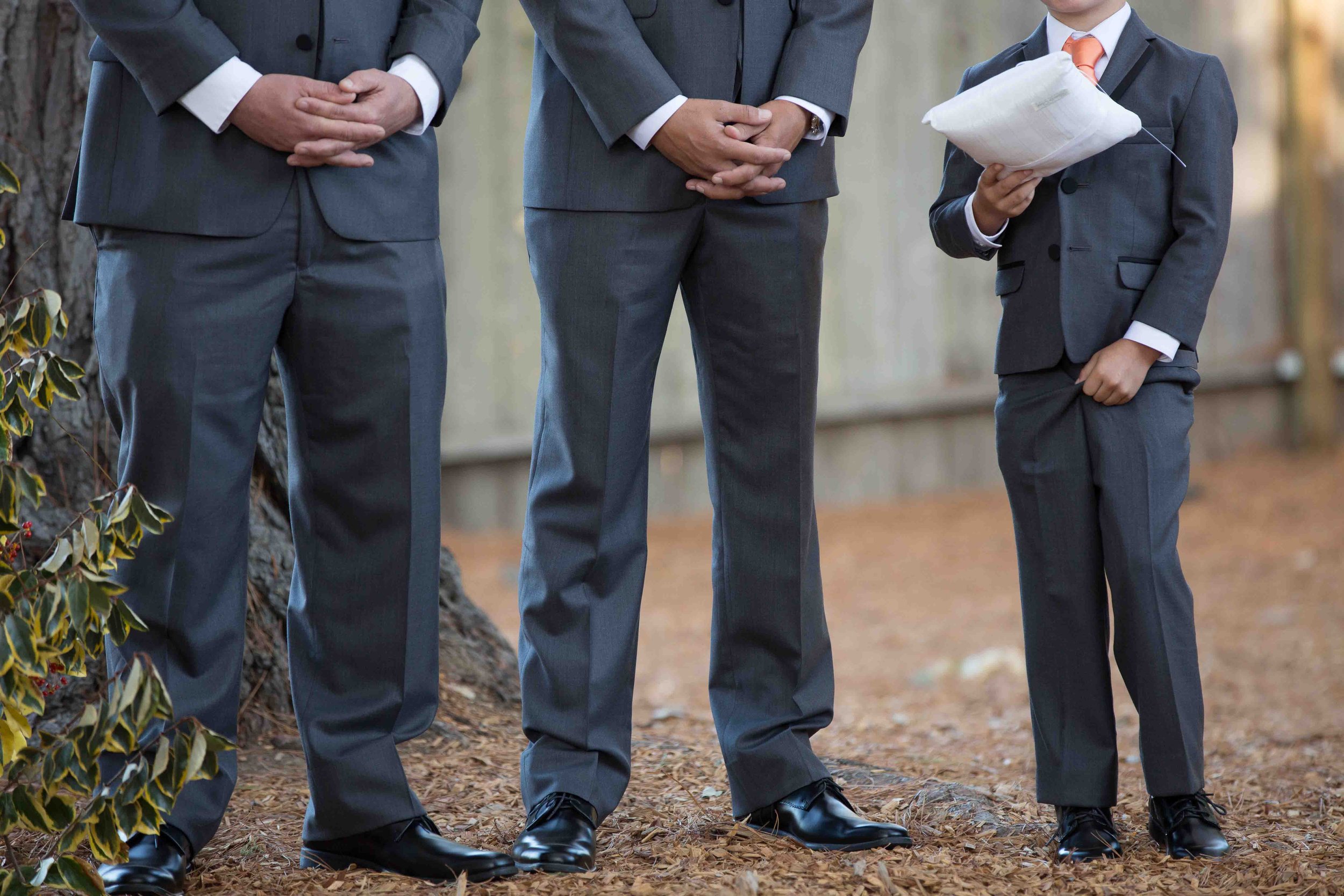 Three people in suits standing outdoors on a dirt ground, with only their lower bodies visible, one of whom is holding a pillow.