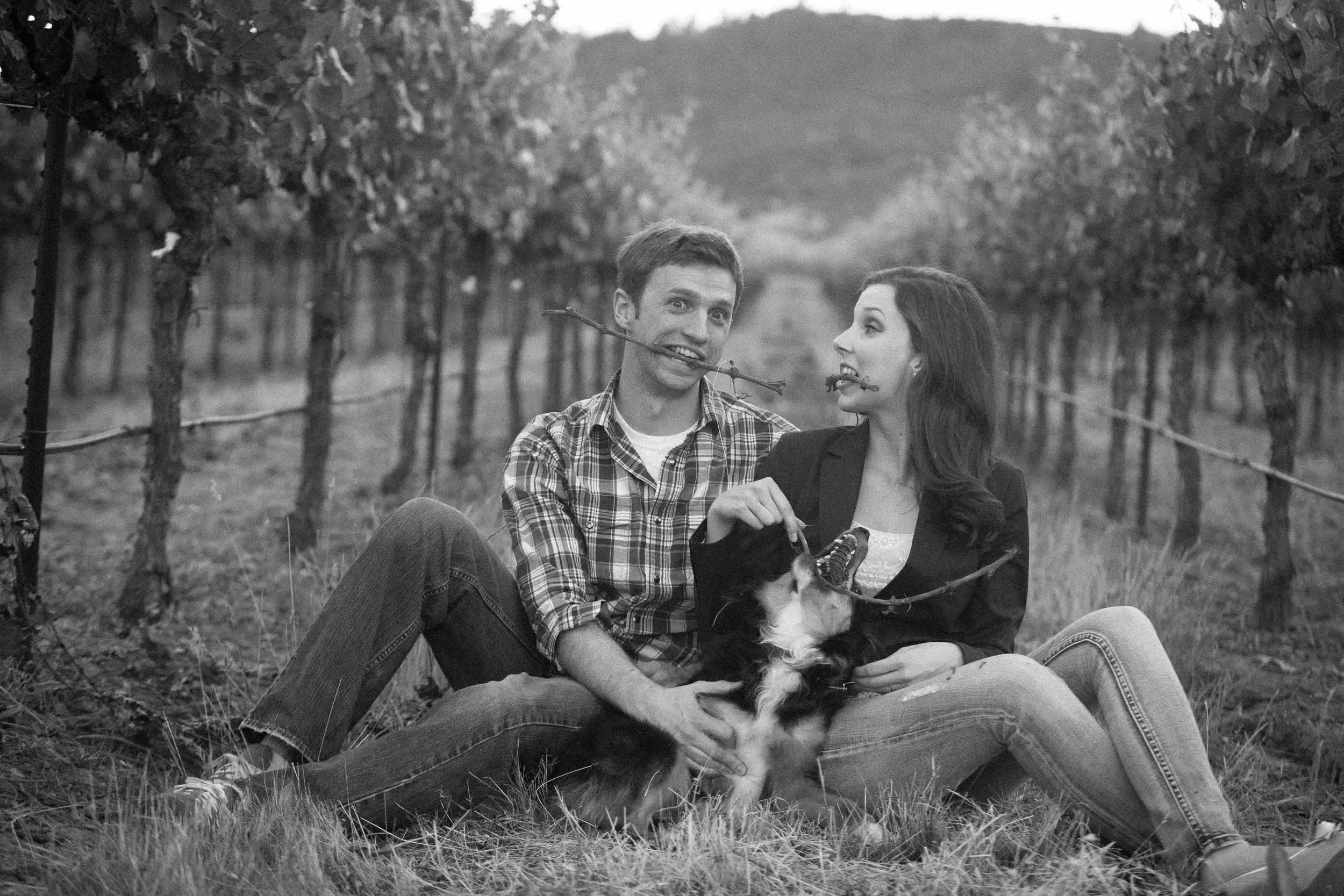 A young couple sitting on the ground in a vineyard, playfully holding sticks in their mouths, with a dog between them. The scene is in black and white.