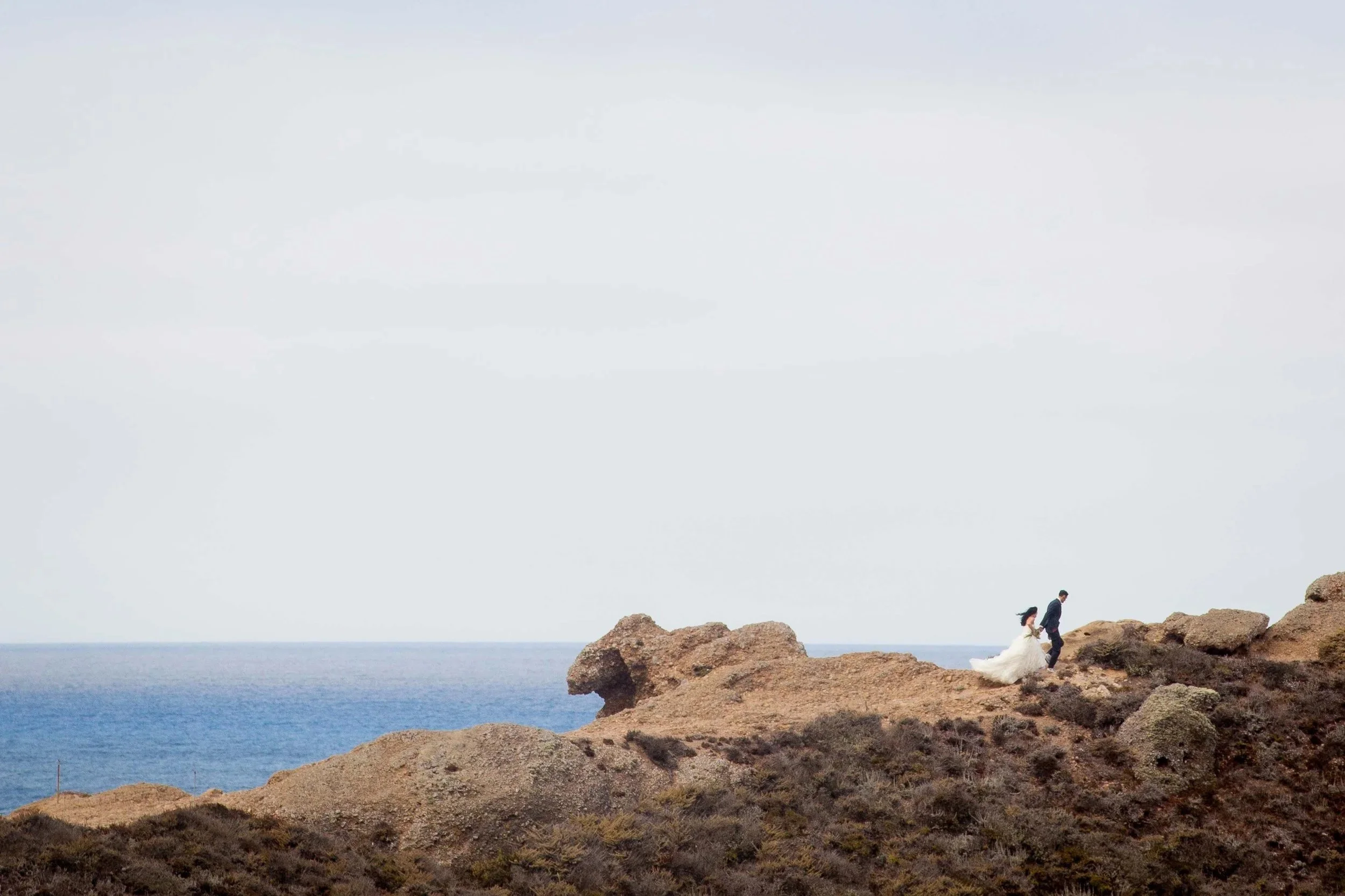 A bride and groom walk on rocky terrain near the ocean, with the bride in a white wedding dress and the groom in a dark suit.
