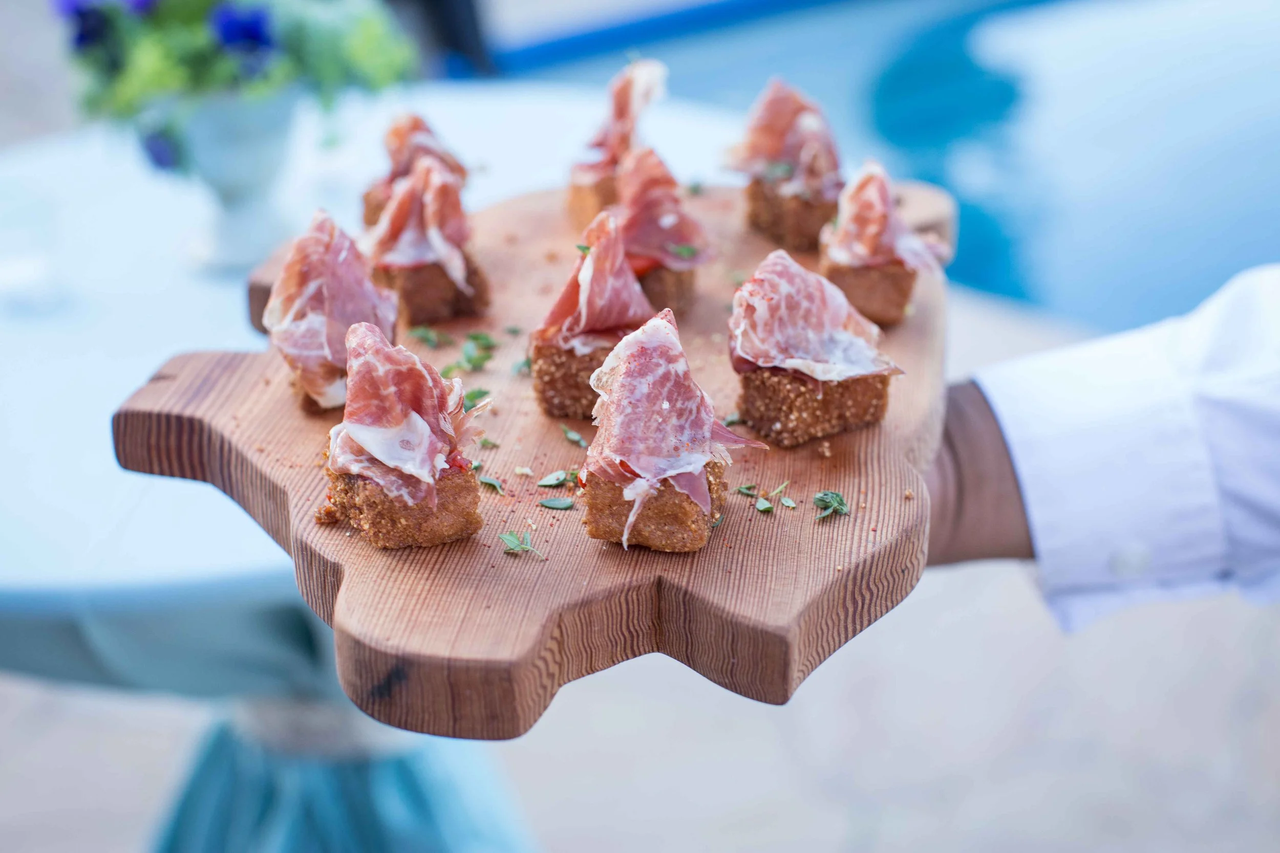 Person holding a wooden serving tray with small bites of food topped with prosciutto. The tray is decorated with small herbs, and the background is blurred.