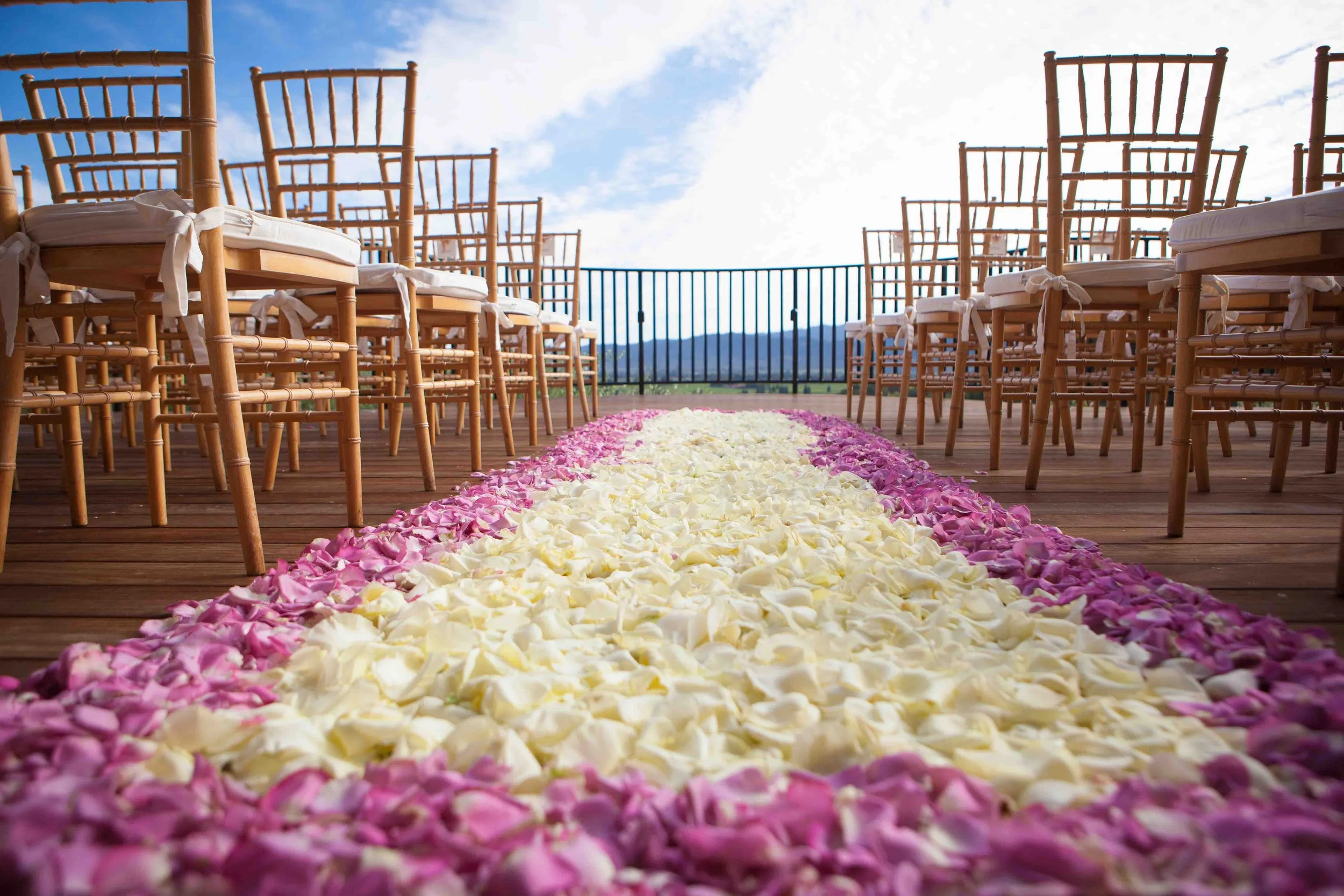 Outdoor wedding aisle decorated with pink and white rose petals on a wooden platform, flanked by chairs with white cushions and bows, overlooking a scenic mountain view.