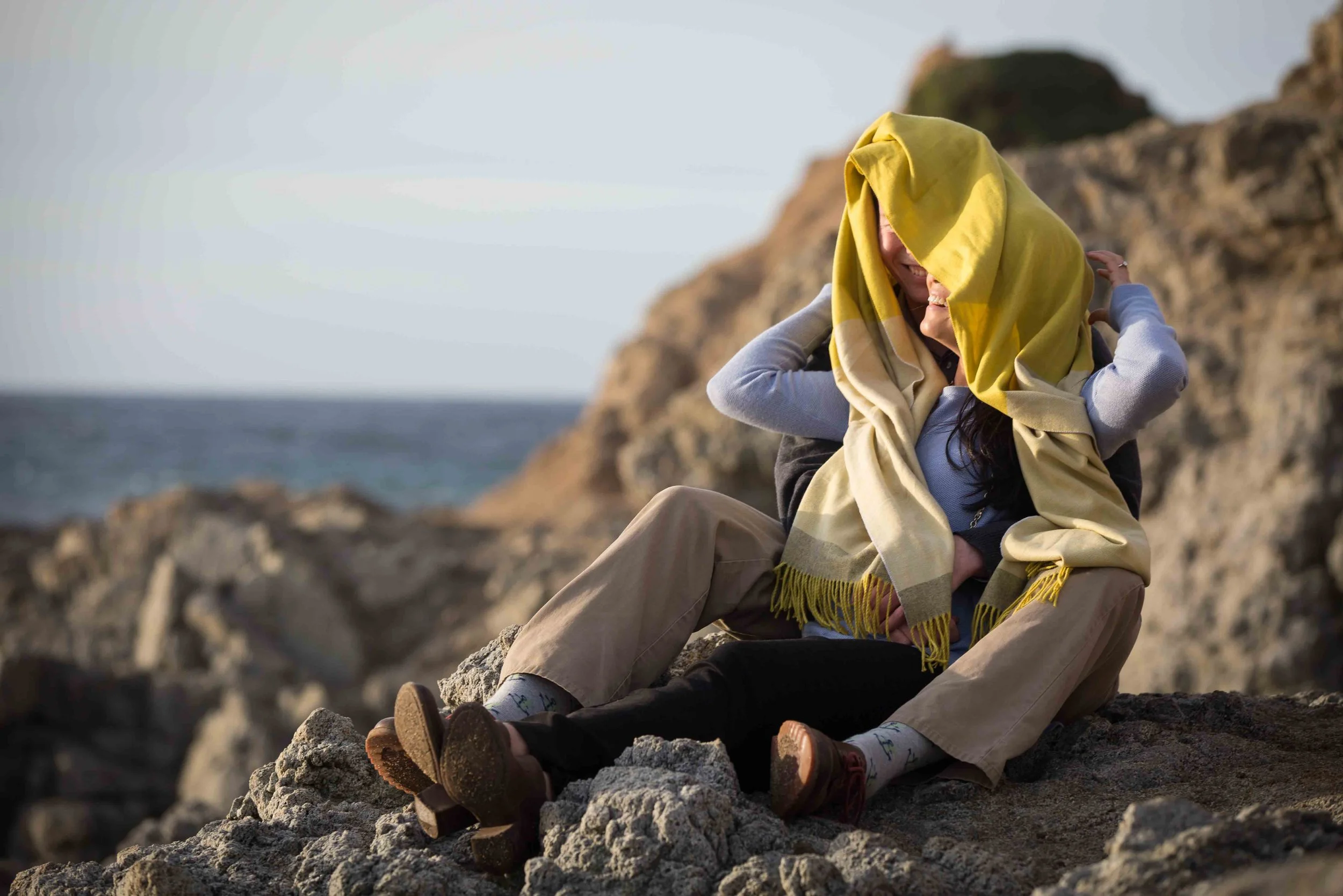 A woman and a girl sitting on rocks at the beach, with the woman covering her face with a yellow scarf, while both are smiling and playing among rocks with the ocean and cliffs in the background.