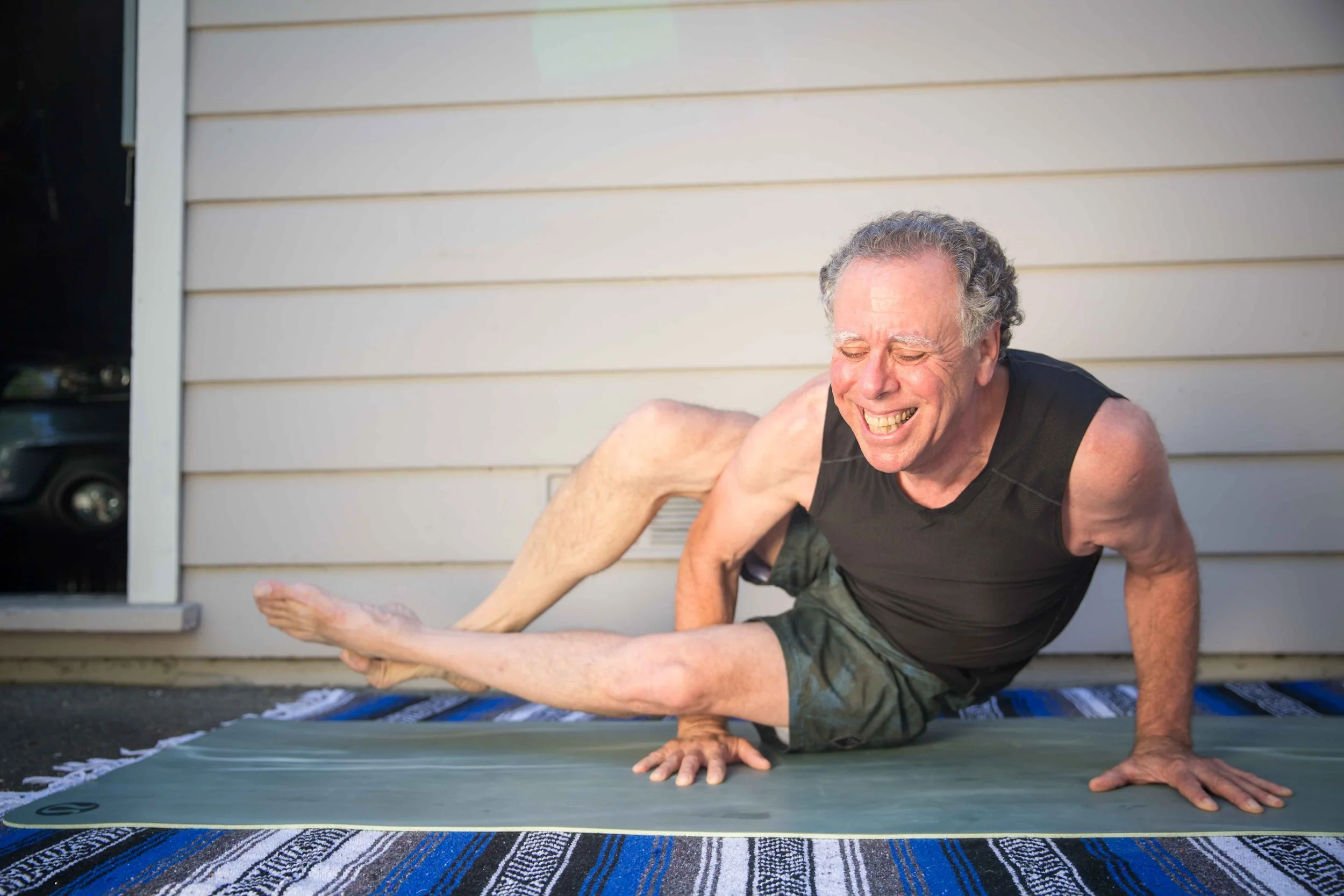 An elderly man with gray hair practicing yoga outside on a mat, smiling with eyes closed, wearing a black sleeveless shirt and camouflage shorts, against a beige house wall.