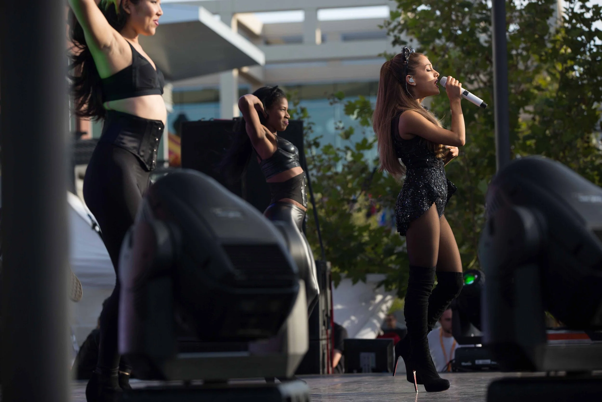 A singer performs on stage with two backup dancers, outdoor setting with trees and modern buildings, singer in black sequin dress and thigh-high boots, backup dancer in black outfit, stage lighting.