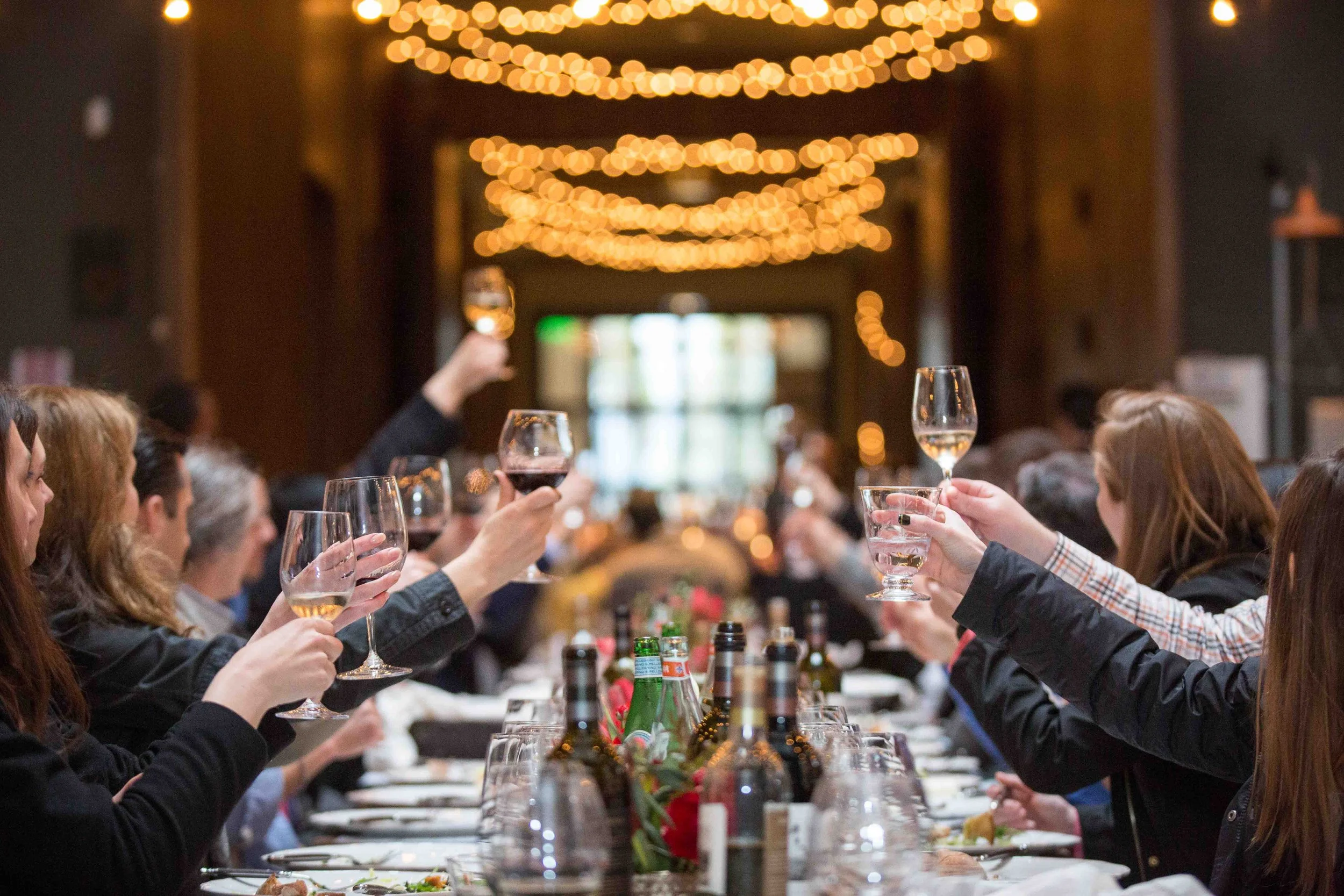 Group of people raising glasses in a toast at a long dinner table decorated with wine bottles and flowers, with warm hanging string lights in the background.