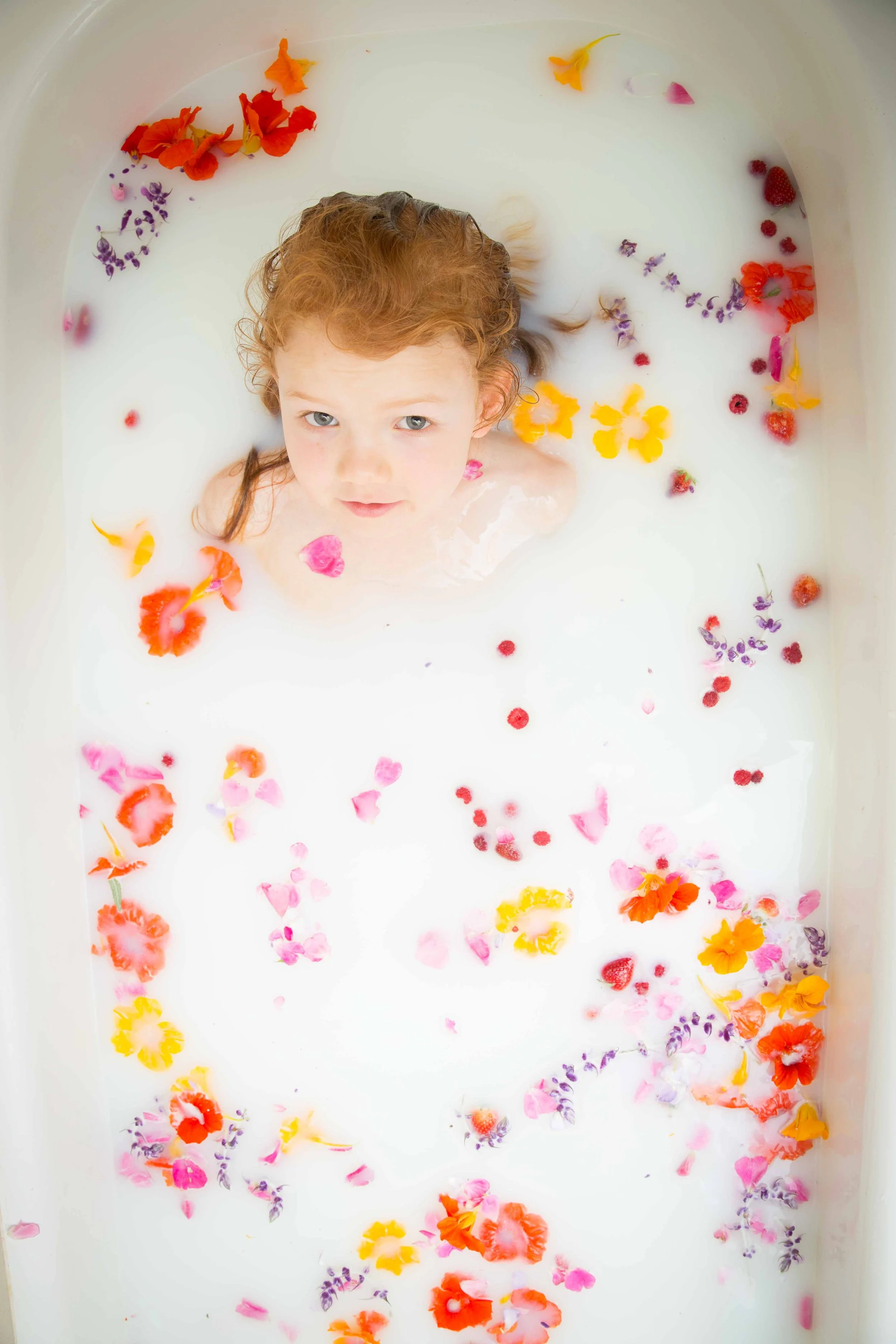 Child with red hair and blue eyes relaxing in a milk bath with colorful flower petals floating around.
