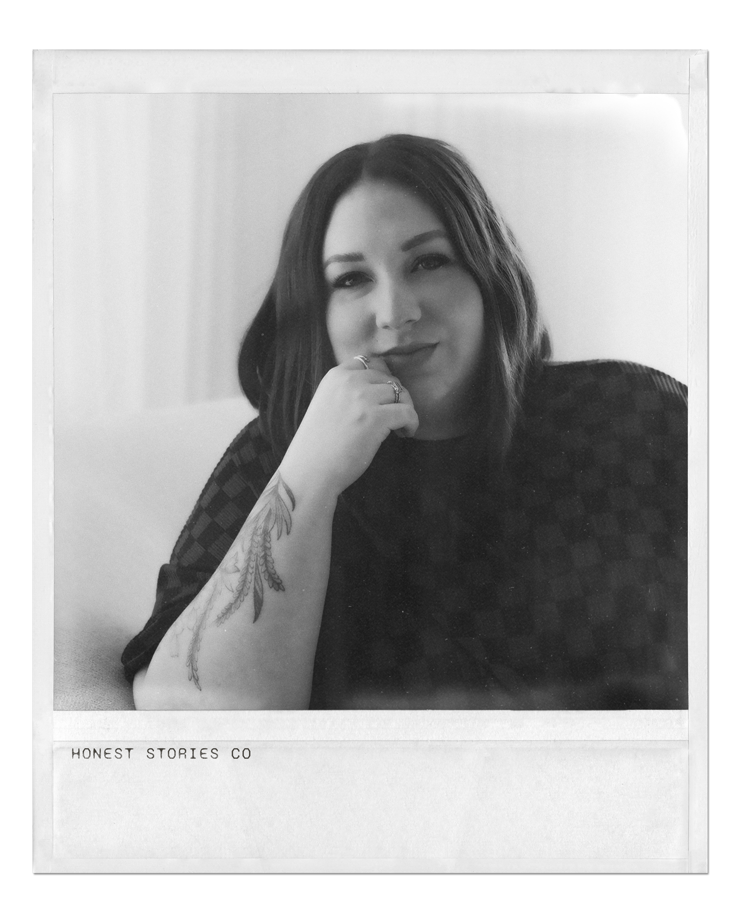 Black and white portrait of a woman with dark hair, wearing a checkered top, resting her chin on her hand, with a tattoo on her forearm and several rings on her fingers. The photo has a vintage Polaroid style with the text 'HONEST STORIES CO' at the bottom.