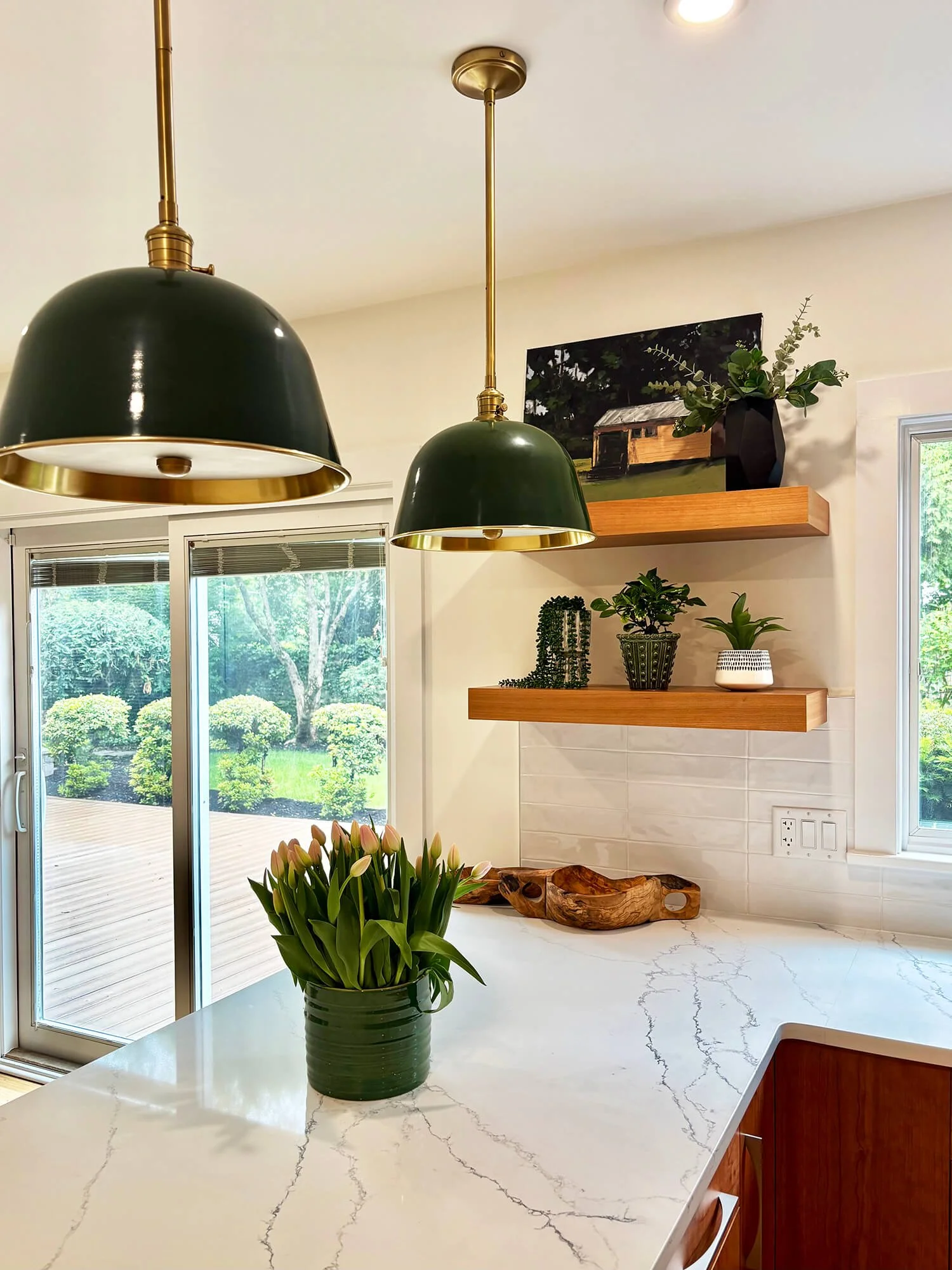 Kitchen with a white marble countertop, a potted plant with pink flowers, and a view of a garden outside through glass sliding doors. Two green pendant lights hang from the ceiling, and wooden shelves with decorative items are on the wall.