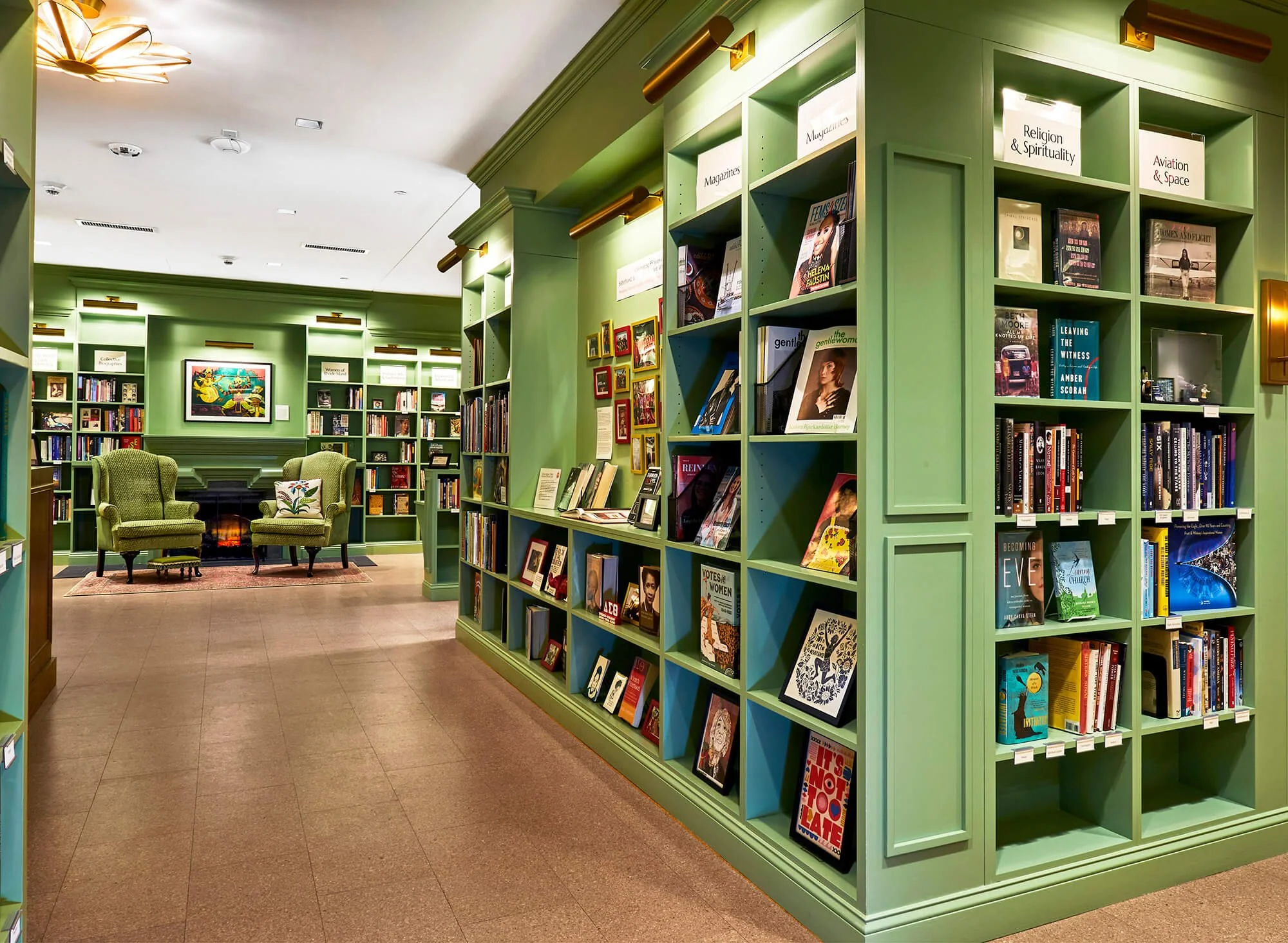 Interior of a bookstore or library with green bookshelves filled with books and magazines, a sitting area with two green armchairs, a small rug, and art on the walls.