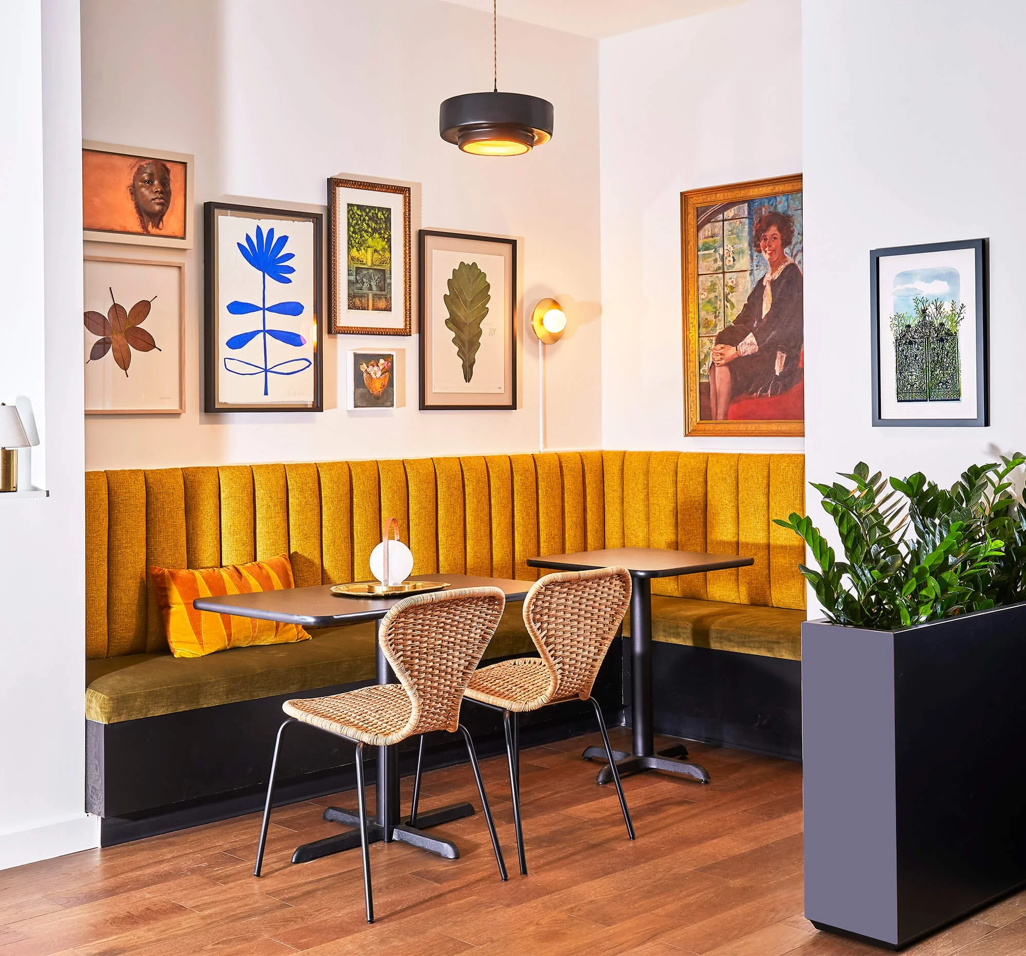 A cozy corner in a restaurant with a mustard yellow upholstered banquette, a small black table, and two woven rattan chairs. The wall behind has various framed artworks, including a blue floral print, a portrait of a woman, a leaf print, and other botanical and nature scenes. There's a modern black ceiling light and a wall-mounted sconce, along with a large green potted plant in the foreground.