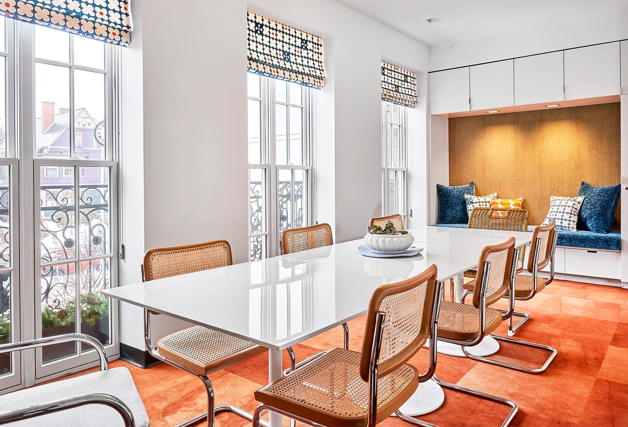 Bright dining room with white walls and large windows with floral patterned Roman shades. White table with wicker chairs around it, a bowl with plants in the center. Built-in white cabinets and a nook with upholstered cushions and pillows on the wall. Orange carpeted floor.