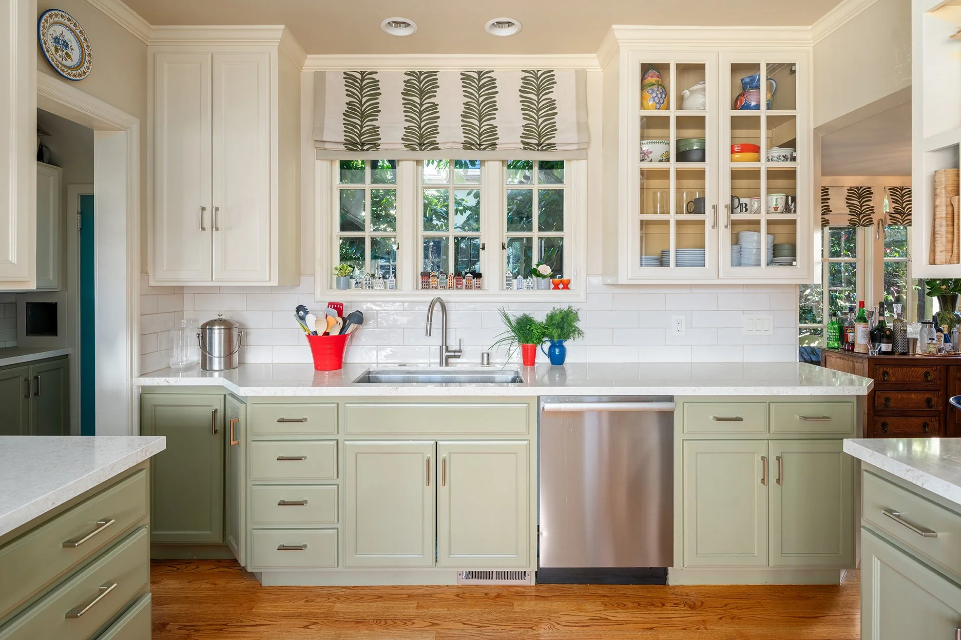 A bright kitchen with white cabinets, a white marble countertop, and a window above the sink with a view of greenery outside. Decorative plants, colorful dishes, and various kitchen tools and bottles are arranged on the countertop and shelves.