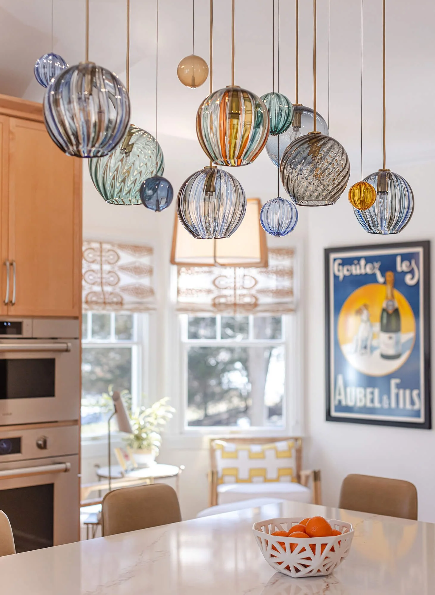 A kitchen or dining area with colorful glass pendant lights hanging from the ceiling, a window with natural light, wooden furniture, a bowl of oranges on a white marble table, and a framed poster on the wall.