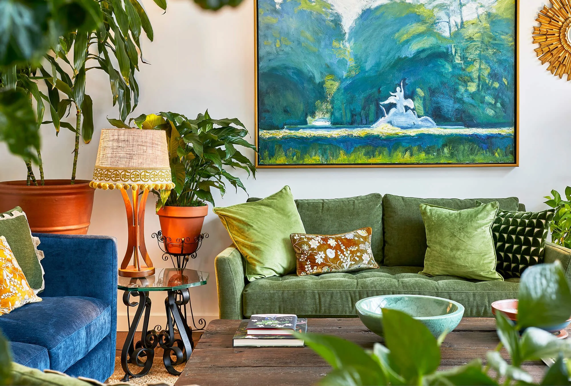 Living room with green velvet sofa and a blue velvet armchair, decorated with green and floral throw pillows. A wooden coffee table with decorative bowls and books is in front. Large potted plants, a table lamp, and a colorful landscape painting on the wall add to the cozy, vibrant ambiance.
