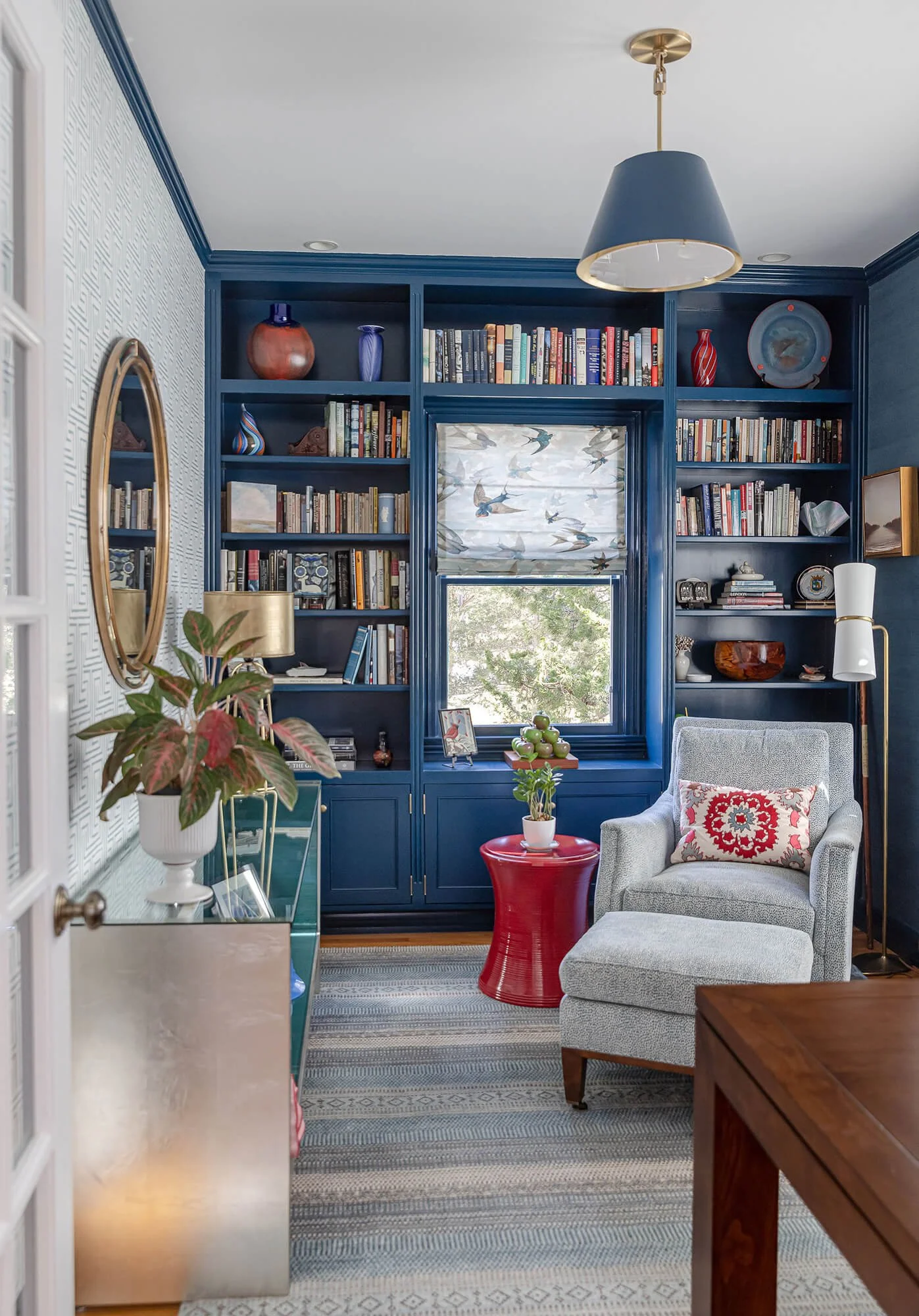 Living room with blue built-in bookshelf filled with books and decorative items, a white armchair with a patterned pillow, a red side table with a potted plant, a window with a bird-themed roman shade, and a ceiling lamp.
