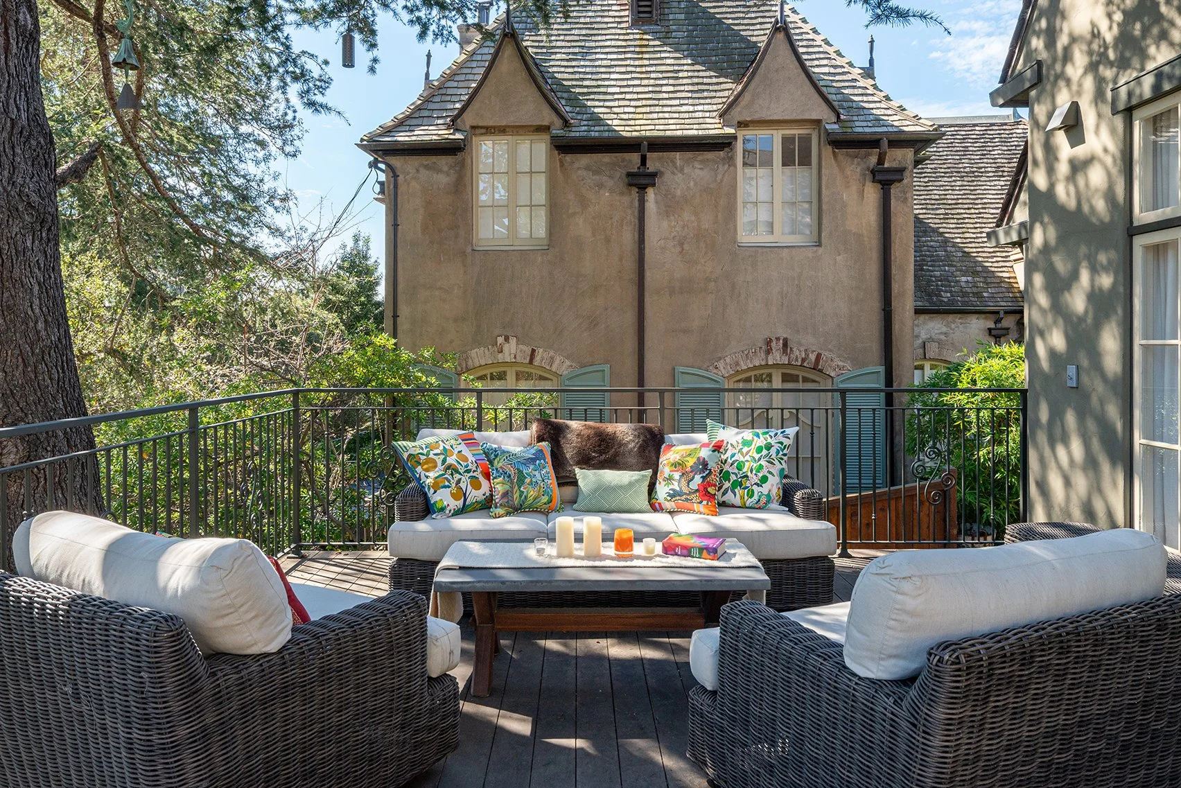 Outdoor patio with wicker seating and colorful pillows, candles, and a table, overlooking trees and a neighboring house with stucco walls, multiple windows, and a steeply pitched roof.