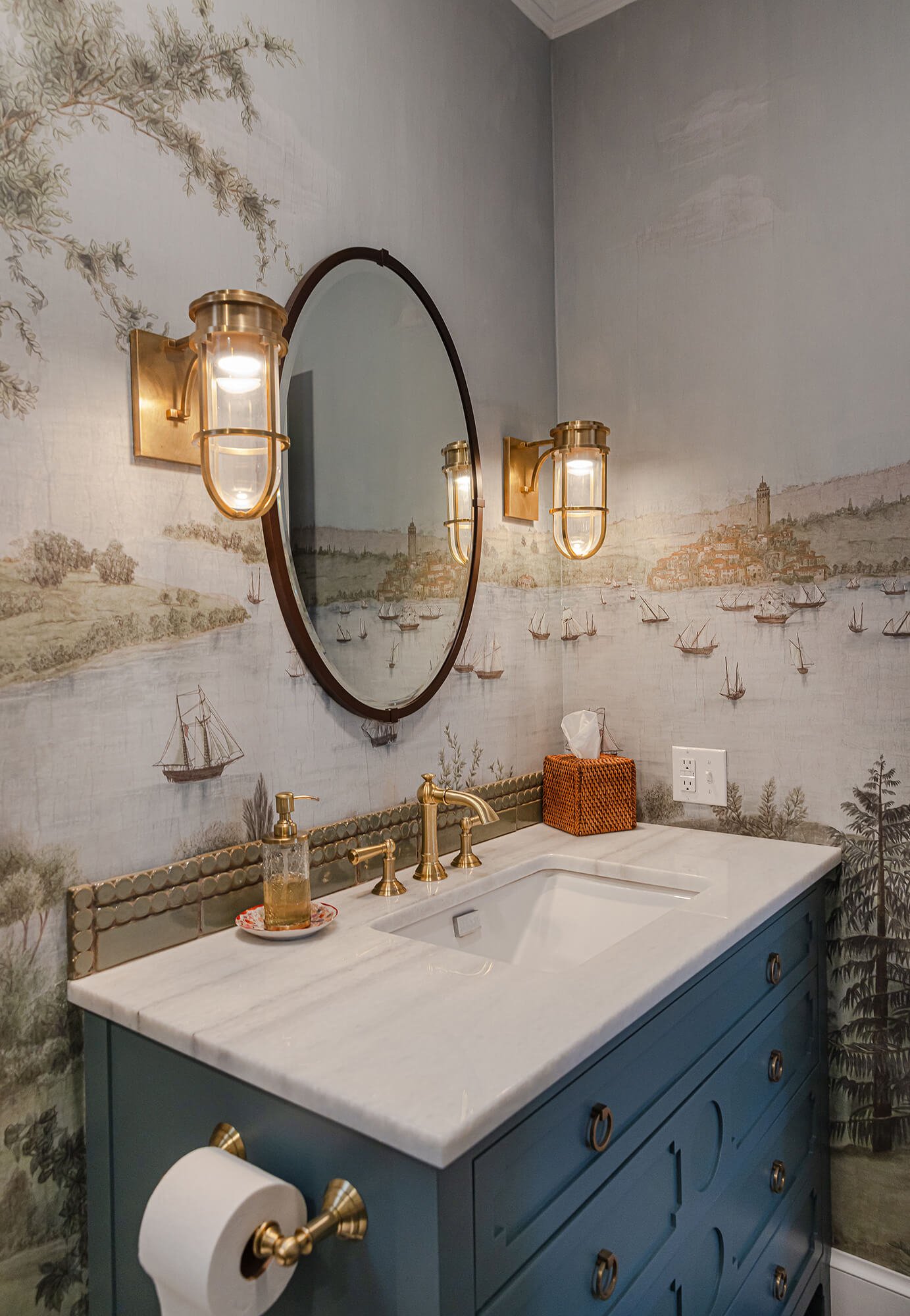Bathroom interior with blue vanity, marble countertop, wall-mounted gold light fixtures, mirror, wallpaper with coastal landscape and sailboats, tissue box, soap dispenser, and toilet paper holder.