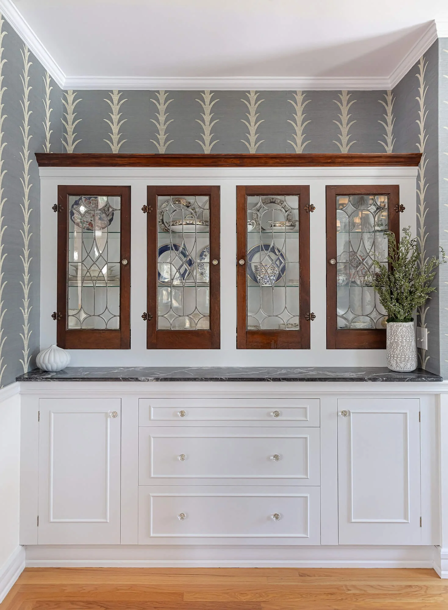 White wooden cabinet with glass doors, decorativeDishware inside, set against a wallpapered wall with a plant in a white planter on the right, and a white decorative object on the left, with wooden flooring and a marble top.