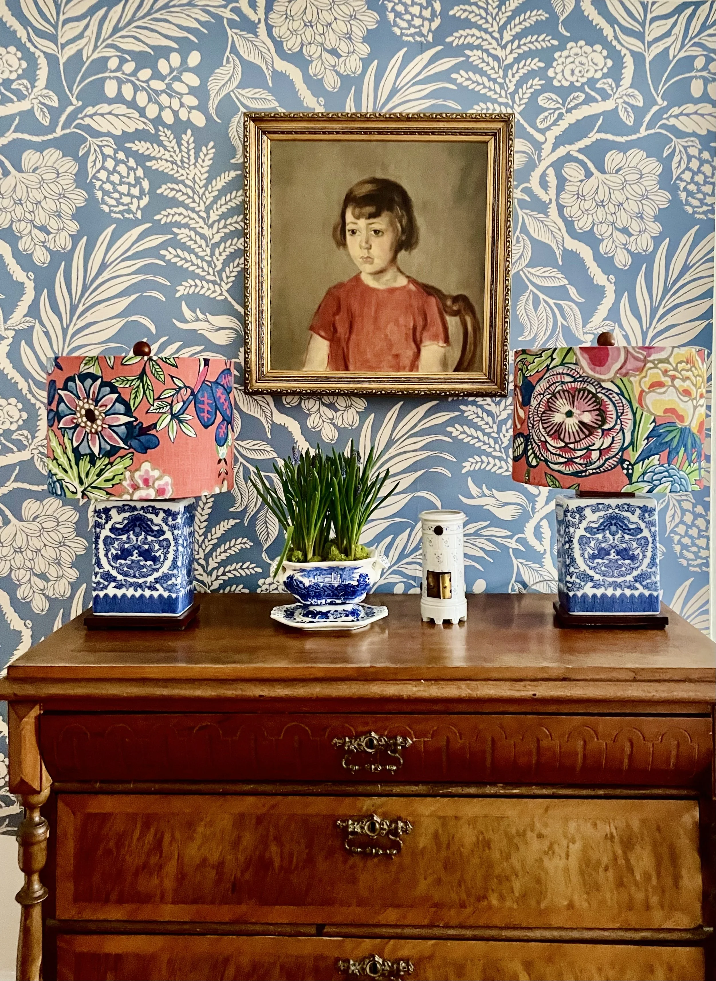 A wooden dresser with two drawers, each with ornate metal handles, behind a table. On the table are two blue and white porcelain lamps with floral lampshades, a potted plant, and a small white decorative container. A framed portrait of a young girl in a red shirt hangs on a blue and white floral patterned wallpaper behind the dresser.