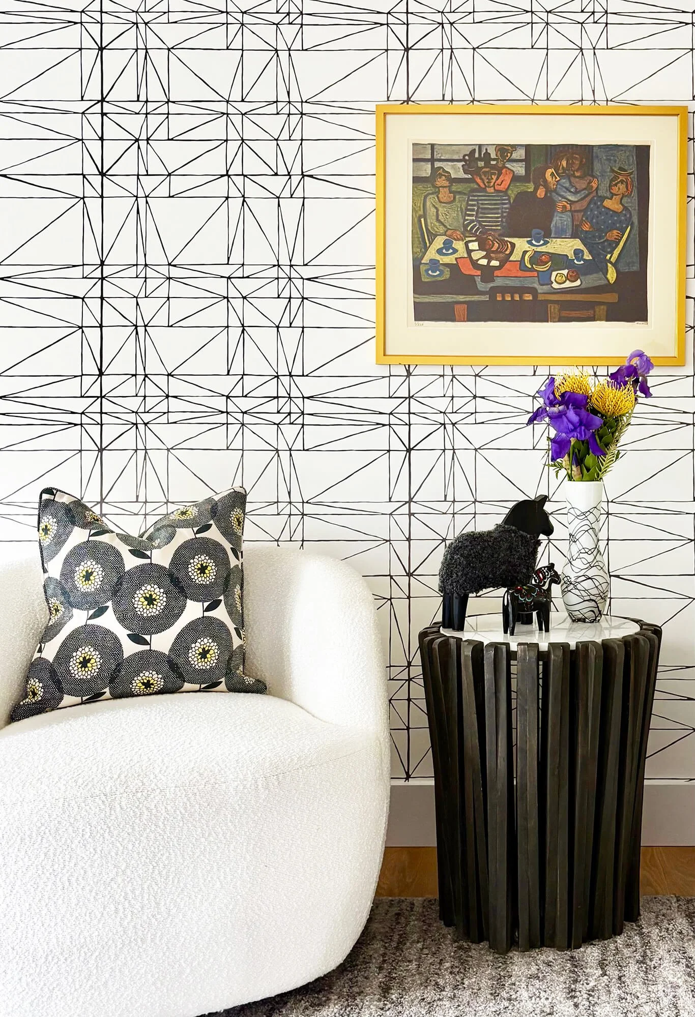 A cozy living room corner featuring a white textured sofa with a black and white floral patterned pillow, a black slatted side table with decorative black animal figurines, a tall white vase with purple irises and yellow flowers, a framed painting of a group of women sitting at a table, and a black and white geometric patterned wall with a gray carpeted floor.
