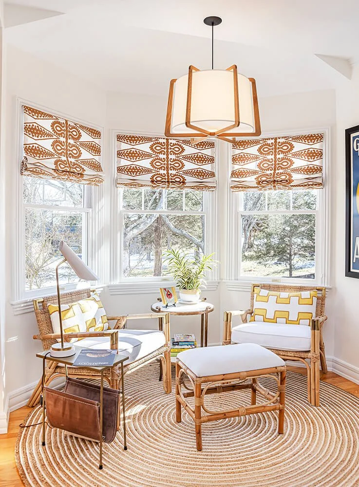 Sunlit sitting area with three rattan chairs with white cushions and yellow patterned pillows, a round woven rug, a small side table with a plant and a framed photo, a floor lamp, and a geometric pendant light, near large windows with decorative blinds showing winter trees outside.