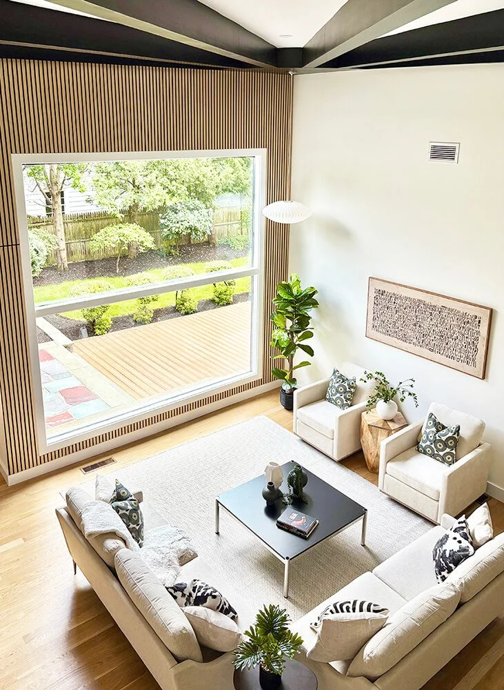 Living room with white sofas, patterned cushions, a black coffee table, potted plants, wall art, and a large window showing a backyard with trees and shrubs.