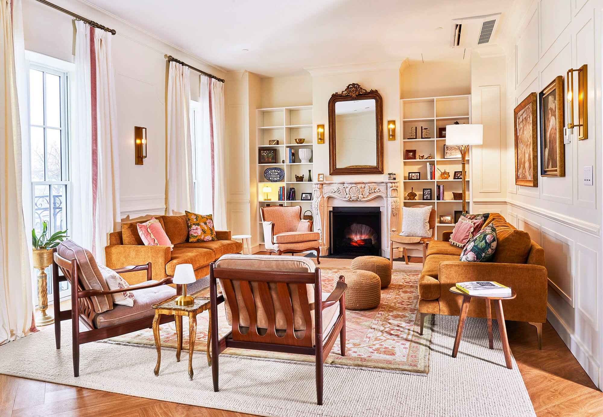 Living room with large windows, white curtains, a stone fireplace with a wooden mirror above, built-in white bookshelves, a mix of vintage and modern furniture, plants, and artwork.