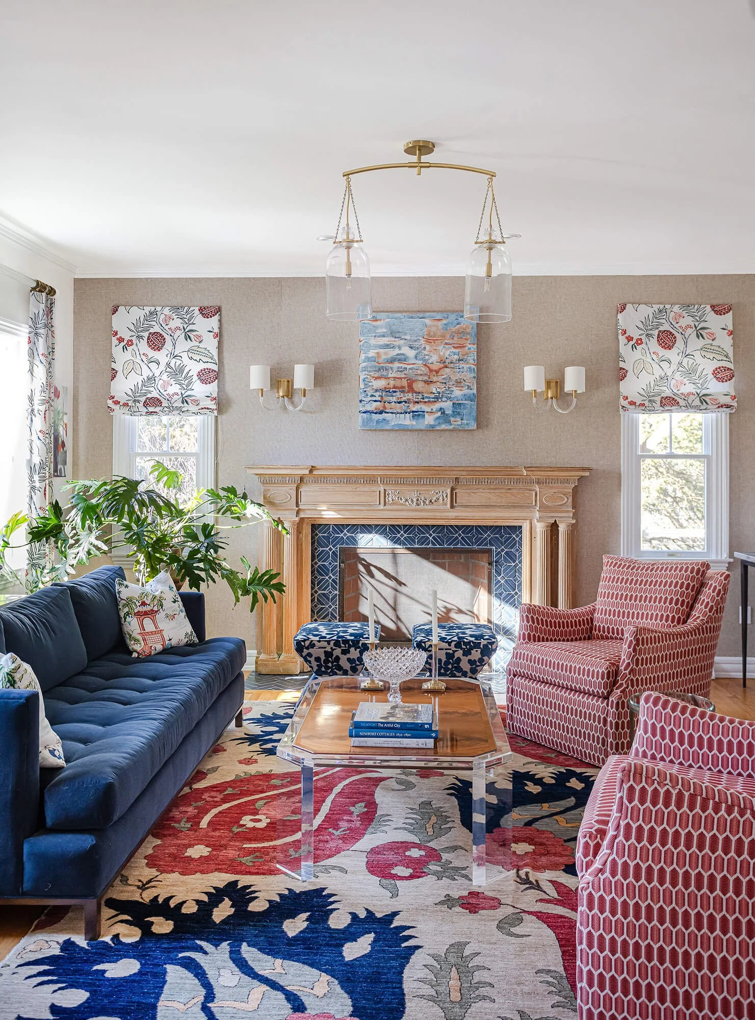 A living room with a fireplace, navy blue velvet sofa, red and white patterned armchair, and a glass coffee table on a floral area rug. There are wall-mounted lamps, floral curtains, and a large plant near the sofa. A modern chandelier hangs from the ceiling, and abstract artwork is above the fireplace.