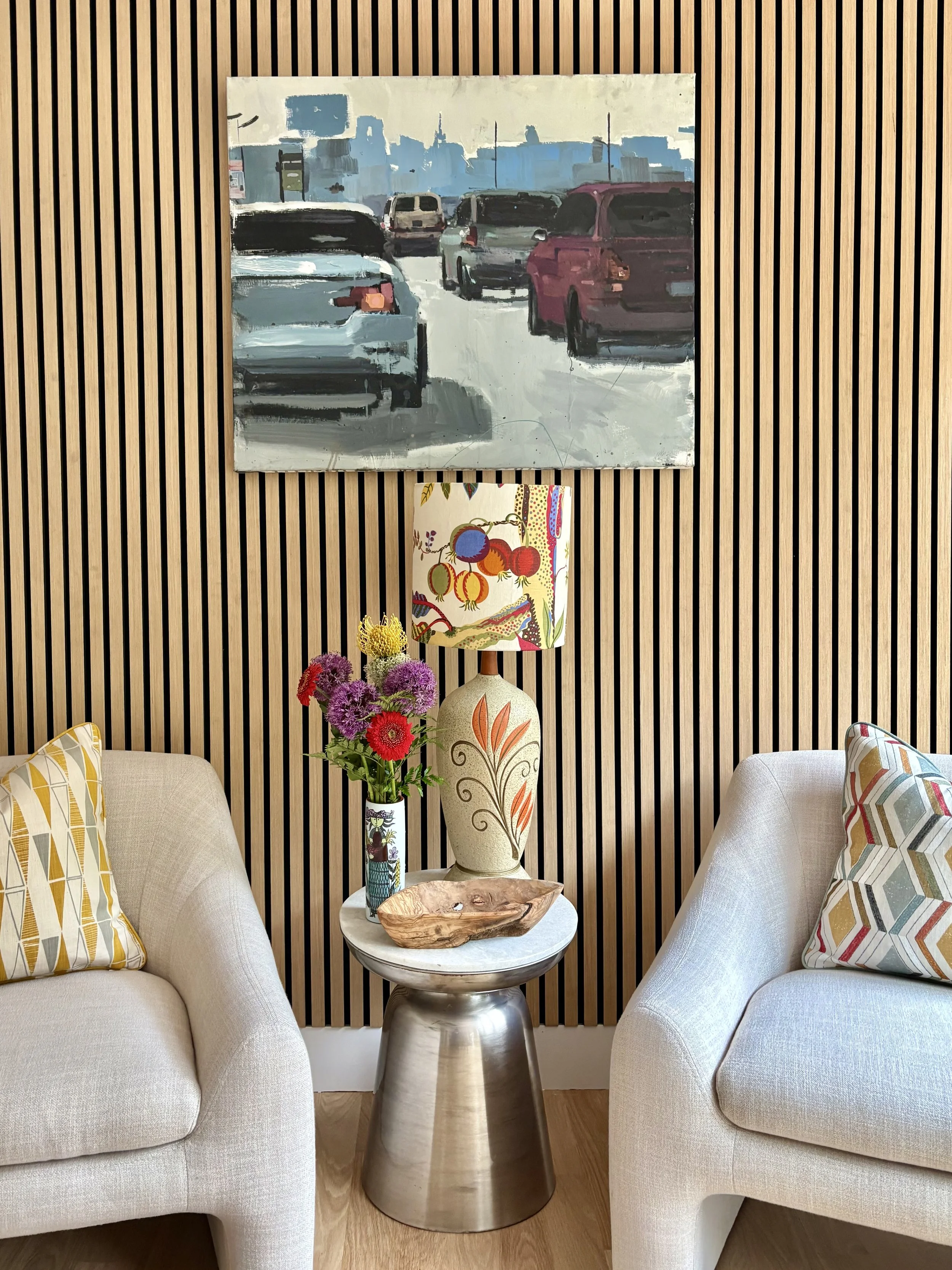 Living room with two beige armchairs, a small round table with a metal base, a tall vase with purple, pink, and yellow flowers, a decorative wooden bowl, a colorful lampshade, and a wooden slat wall with a cityscape painting hanging above the table.