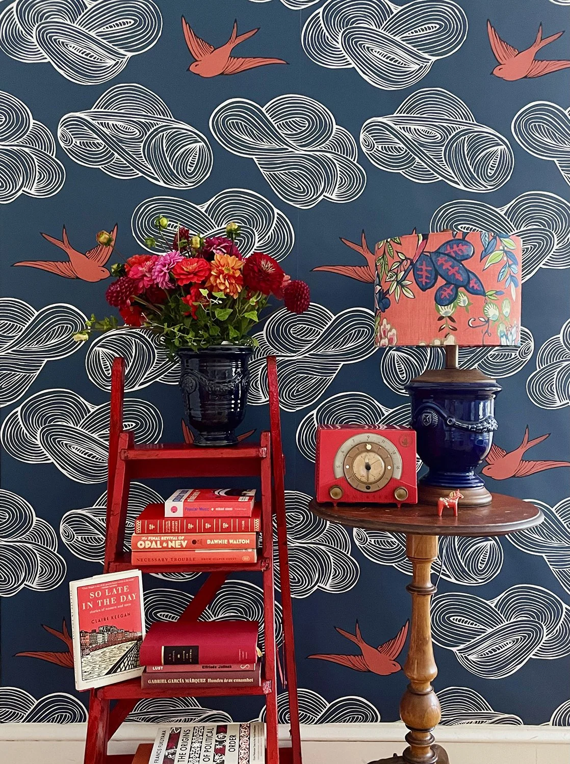 A colorful interior scene with a red ladder holding a black vase filled with a vibrant pink, red, and orange flower arrangement, next to a small table with a blue ceramic lamp with a floral lampshade, a red vintage clock, and a small pink donkey figurine. The background features a blue wallpaper with white swirling cloud patterns and red flying bird illustrations.