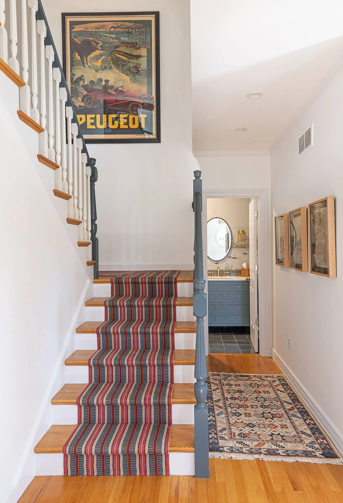 Interior view of a house entryway showing a staircase with a striped runner, a hallway with a patterned rug, framed artwork, a doorway to a bathroom with a vanity and mirror, and a overhanging ceiling with recessed lighting.