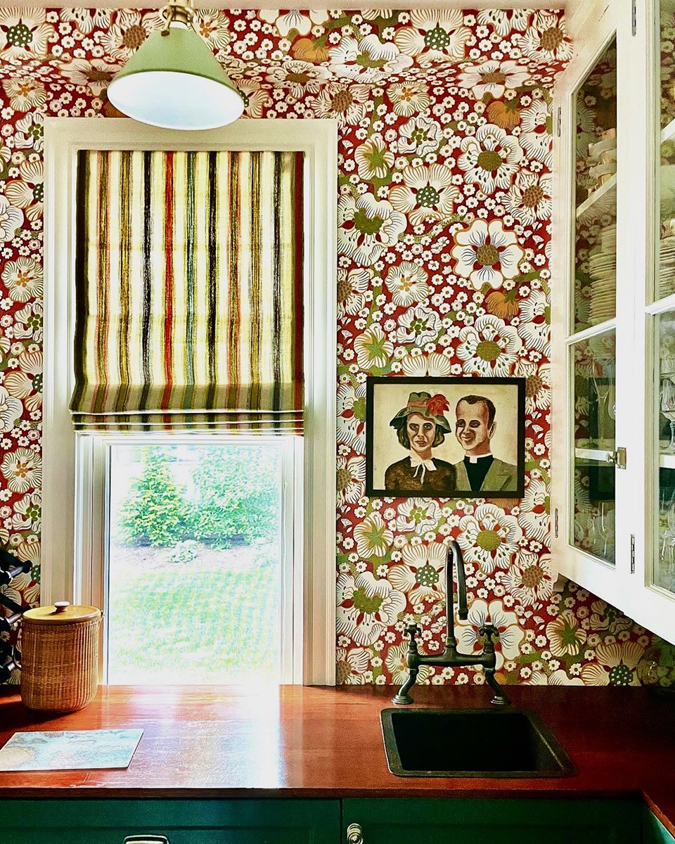 Kitchen with floral wallpaper, a window with striped Roman shade, a painting of a man and woman, a faucet, and a wicker container.
