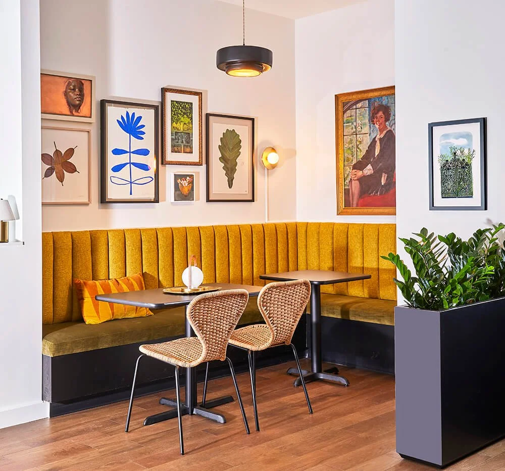 A cozy café corner with a yellow upholstered banquette, two woven rattan chairs, and framed art on white walls, including portraits, foliage, and abstract images. There's a black pendant light, a wall sconce, a small table lamp on the table, and a large potted plant.