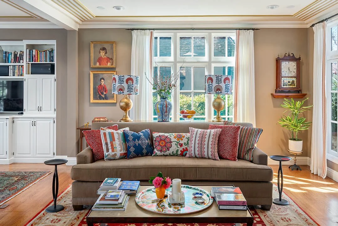 Cozy living room with a beige sofa adorned with colorful and patterned throw pillows. A coffee table with books, a vase, and candles is in front. Behind the sofa, a large window with white curtains, flanked by two table lamps with decorative shades, lets in natural light. To the left, a white built-in bookshelf with books and a television. To the right, a wall clock and a potted plant, with artwork on the walls.