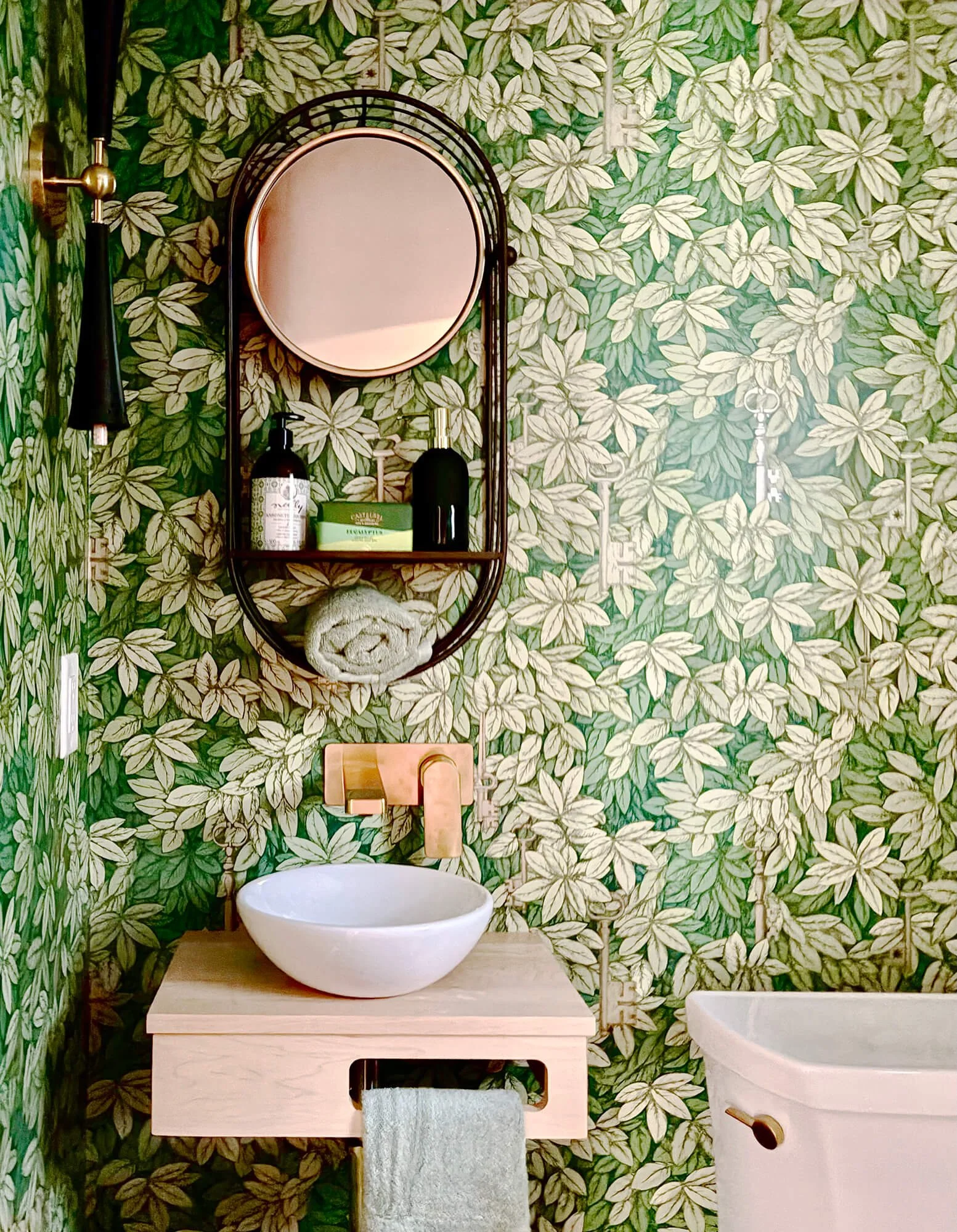 Bathroom with green and cream leaf patterned wallpaper. Features a round mirror, a wooden vanity with a white vessel sink, a towel hanging underneath, a soap dispenser, a green box, and a hairdryer. There is a small white bathtub on the right.