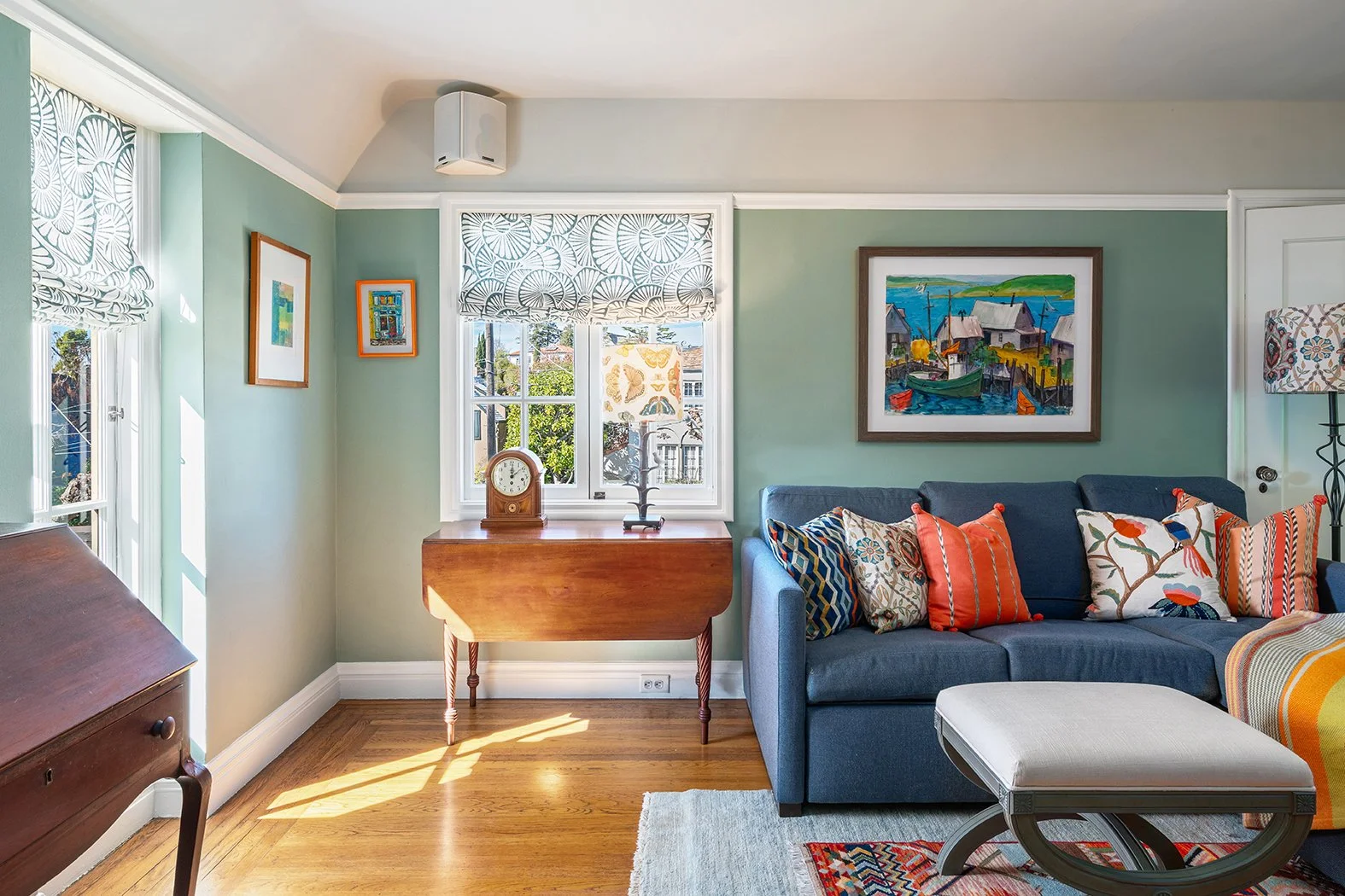 Living room with green walls, wooden floor, and a gray couch adorned with colorful patterned pillows. There is a wooden table beneath a window with a clock and a decorative object. Framed artwork hangs on the walls, and a floor lamp with a patterned shade is beside the couch.