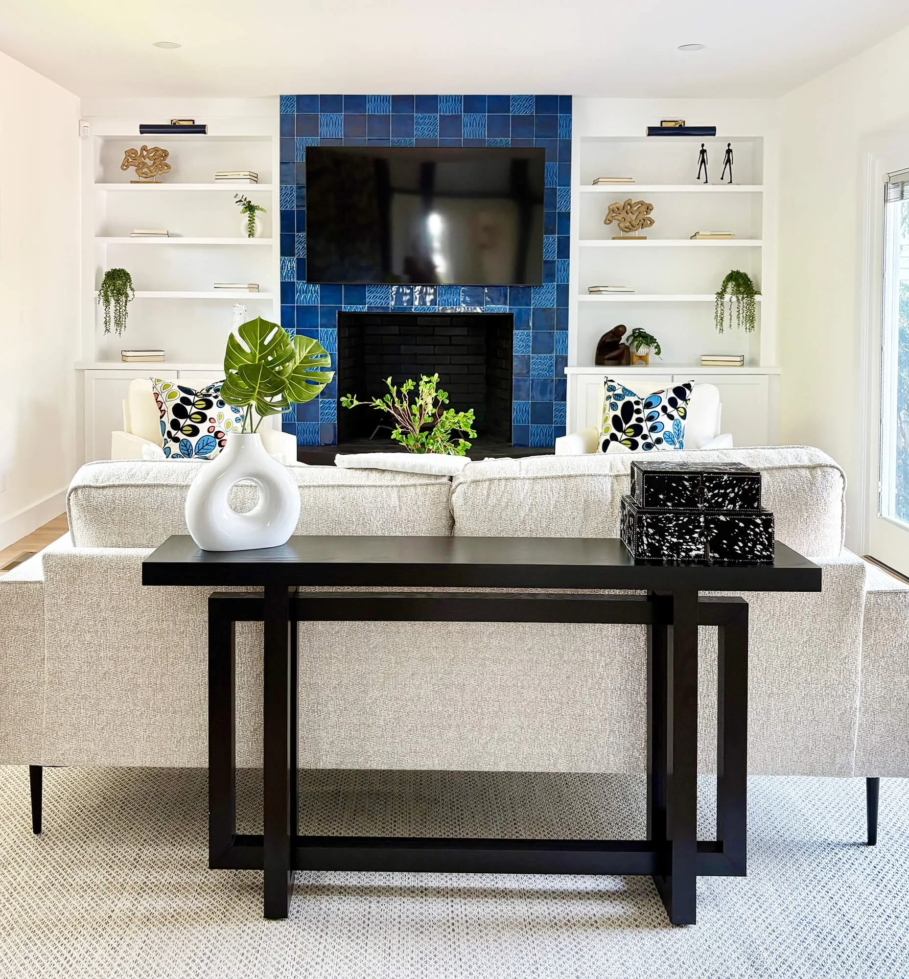 Living room with a black fireplace surrounded by blue tiles, white built-in shelves decorated with books, plants, and sculptures, a wall-mounted TV, a white couch with black, yellow, blue, and white patterned pillows, black decorative boxes, a black coffee table, and a beige carpet.