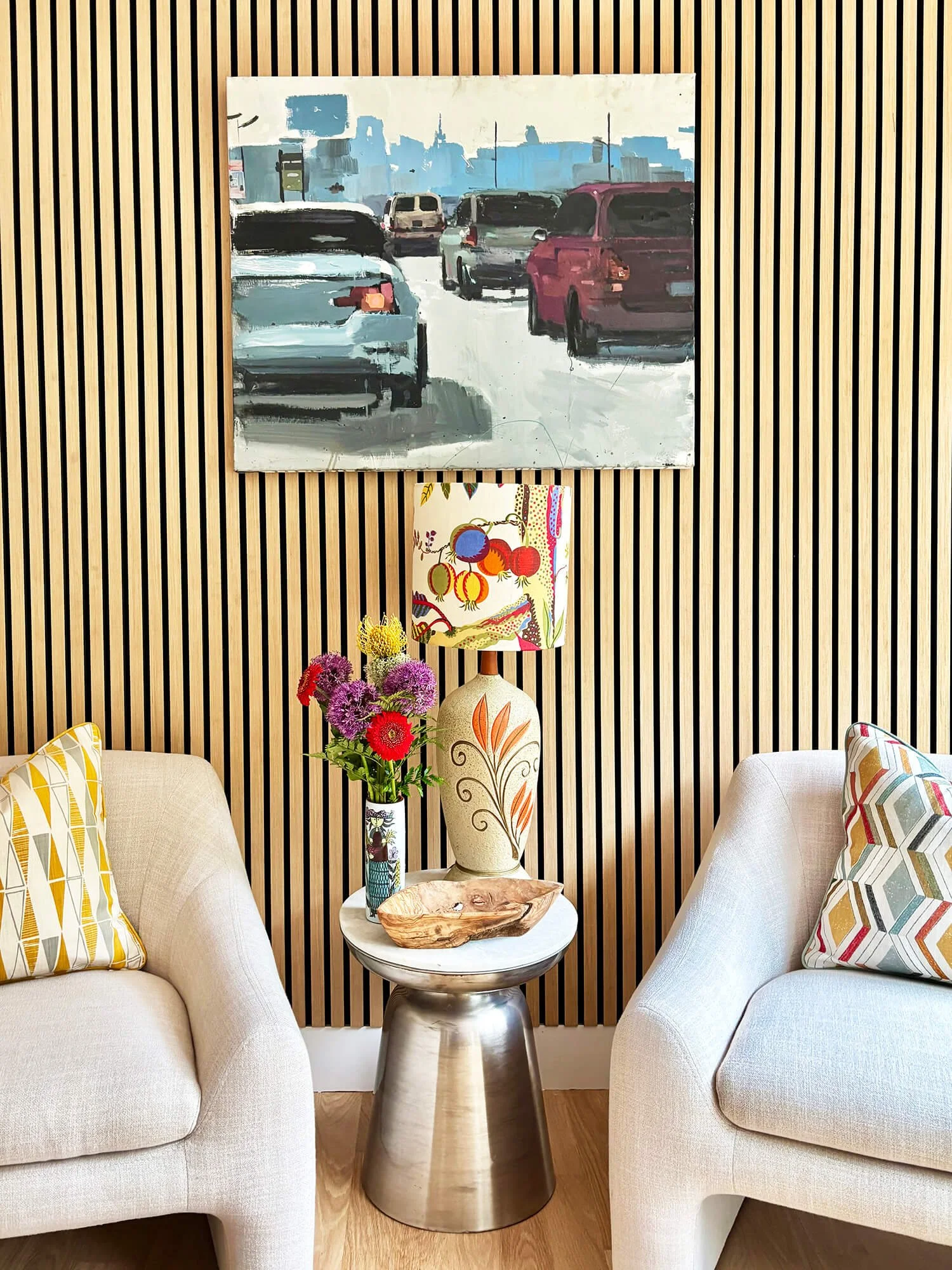 Living room with two cream-colored couches, colorful pillows, a small round white table, a metallic side table, a large decorative vase with flowers, a colorful table lamp, a wooden tray, a painting of cars on a city street, and a wood-paneled wall.