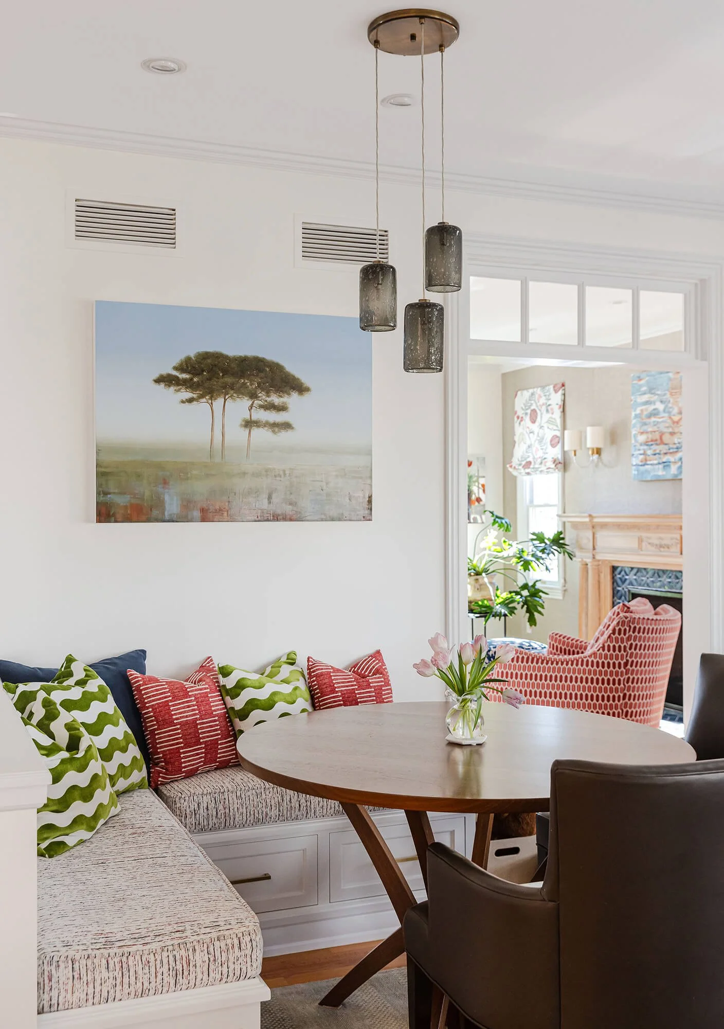 A cozy dining area with a round wooden table, a vase of pink flowers, colorful patterned pillows on a built-in sofa, and a red and white patterned armchair. The room has a large window, a fireplace, and decorative wall art, with modern pendant lighting hanging from the ceiling.