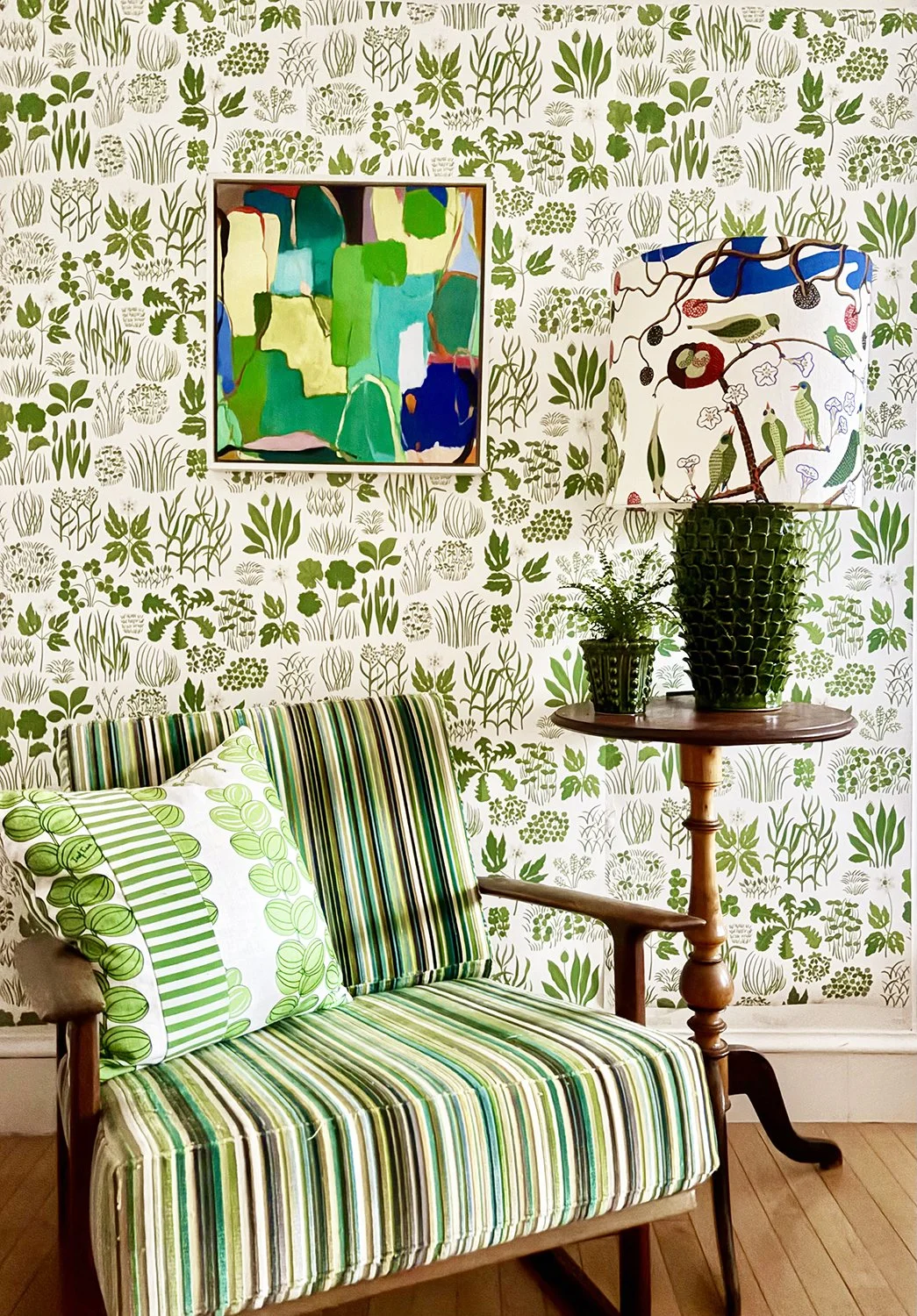 A room with green botanical wallpaper, a colorful abstract painting, a striped green and white upholstered wooden armchair with matching pillows, and a wooden side table holding two large green potted plants and a table lamp with a floral patterned shade.